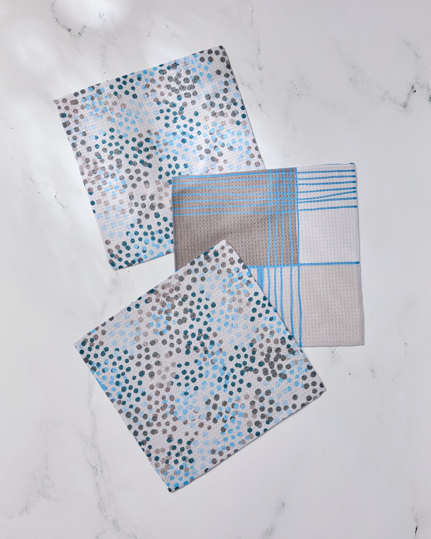 Three Geometry dishcloths from the Cool Blocks and Dots set are displayed on a white marble surface—two featuring blue, grey, and black dot patterns, and one with a blue plaid design—all made from recycled materials.