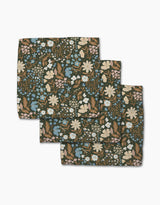Three folded Cottage Meadow Sepia cloth napkins from Danelys Sidron, made of Geoweave fabric with a floral print in white, blue, and beige flowers plus green leaves on a dark background, are arranged in a layered stack.