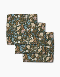 Three folded Cottage Meadow Sepia cloth napkins from Danelys Sidron, made of Geoweave fabric with a floral print in white, blue, and beige flowers plus green leaves on a dark background, are arranged in a layered stack.
