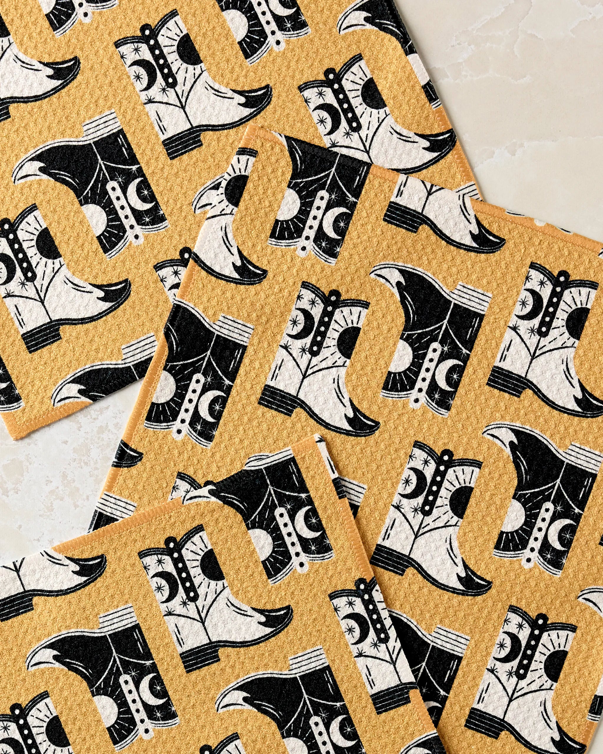 The Megan Roy "Cowboy Boots" set includes four yellow, super-absorbent dishcloths with a black and white cowboy boot pattern—stylish and practical for any kitchen.