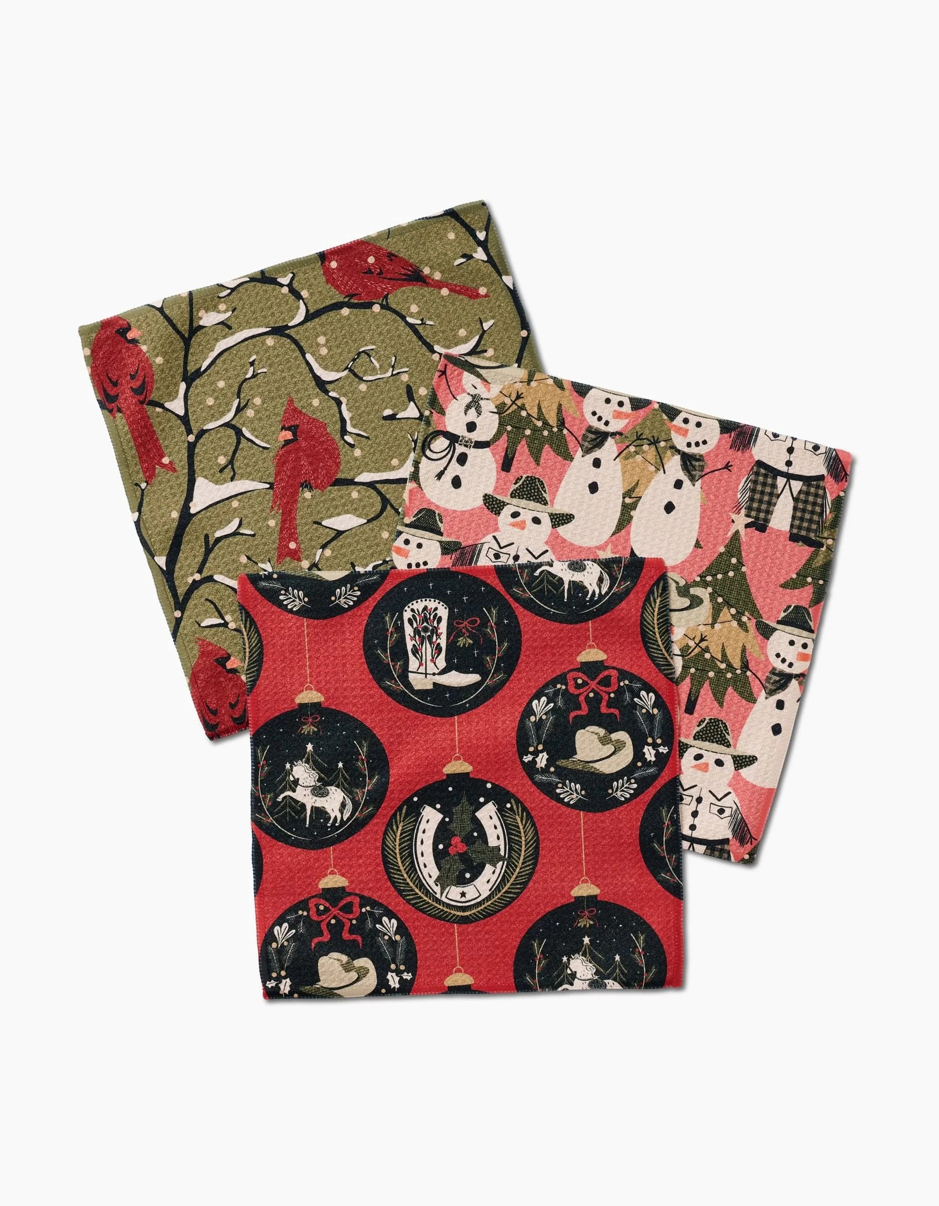 The Cowboy Winter dishcloth set by Julianne Haness includes three recycled cloth napkins featuring red cardinals on branches, snowmen with holiday trees, and Christmas ornaments with reindeer, trees, and gingerbread men.