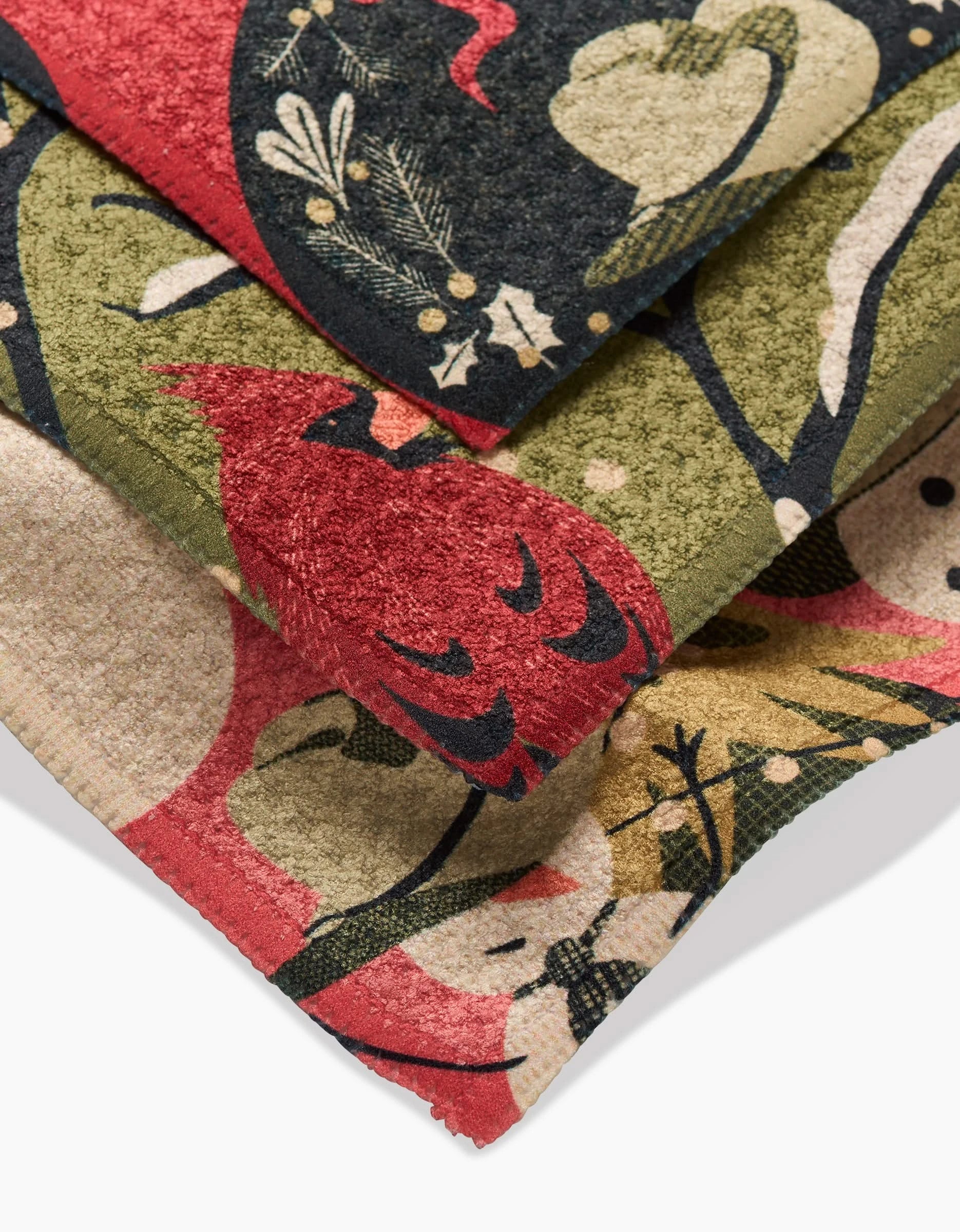 A close-up of three folded Cowboy Winter towels by Julianne Haness, made from recycled materials and featuring a festive red, green, and beige pattern with cardinals, hearts, and branches.
