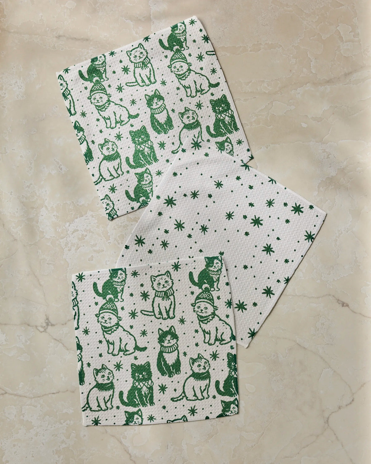 Three white cloths with green prints lie on a beige marble surface. The Geometry Cozy Kittens super-absorbent dishcloth set includes two cloths featuring whimsical cats in hats and scarves, while the third displays green stars and snowflakes.
