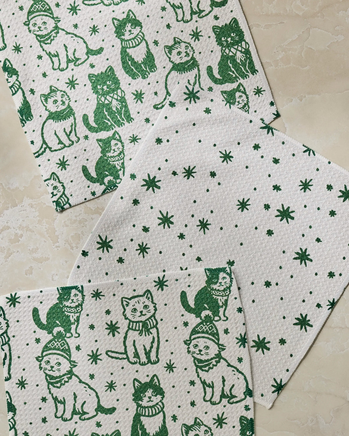 Three white cloths from Geometry’s Cozy Kittens set rest on a beige surface: two feature green illustrations of sweater- and hat-clad cats, while the third displays a playful green star and dot pattern. Crafted from recycled materials, these super-absorbent dish towels are both practical and delightfully charming.