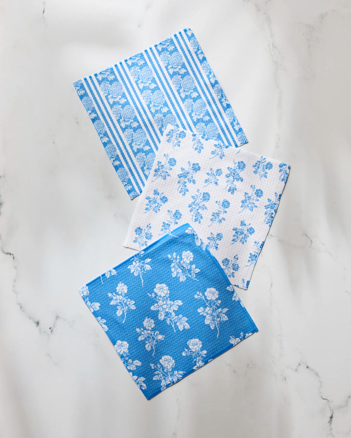 Three Crete square paper napkins by Geometry, featuring blue and white floral patterns, are displayed on a white marble surface. This elegant set complements any table and pairs perfectly with the matching Crete dishcloth set.