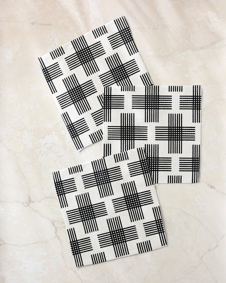 Three square Cross Hatch napkins by Rosa Kichline, made from recycled materials and featuring a black intersecting line pattern, are arranged diagonally with slight overlap on a light marble surface for a modern, geometric effect.