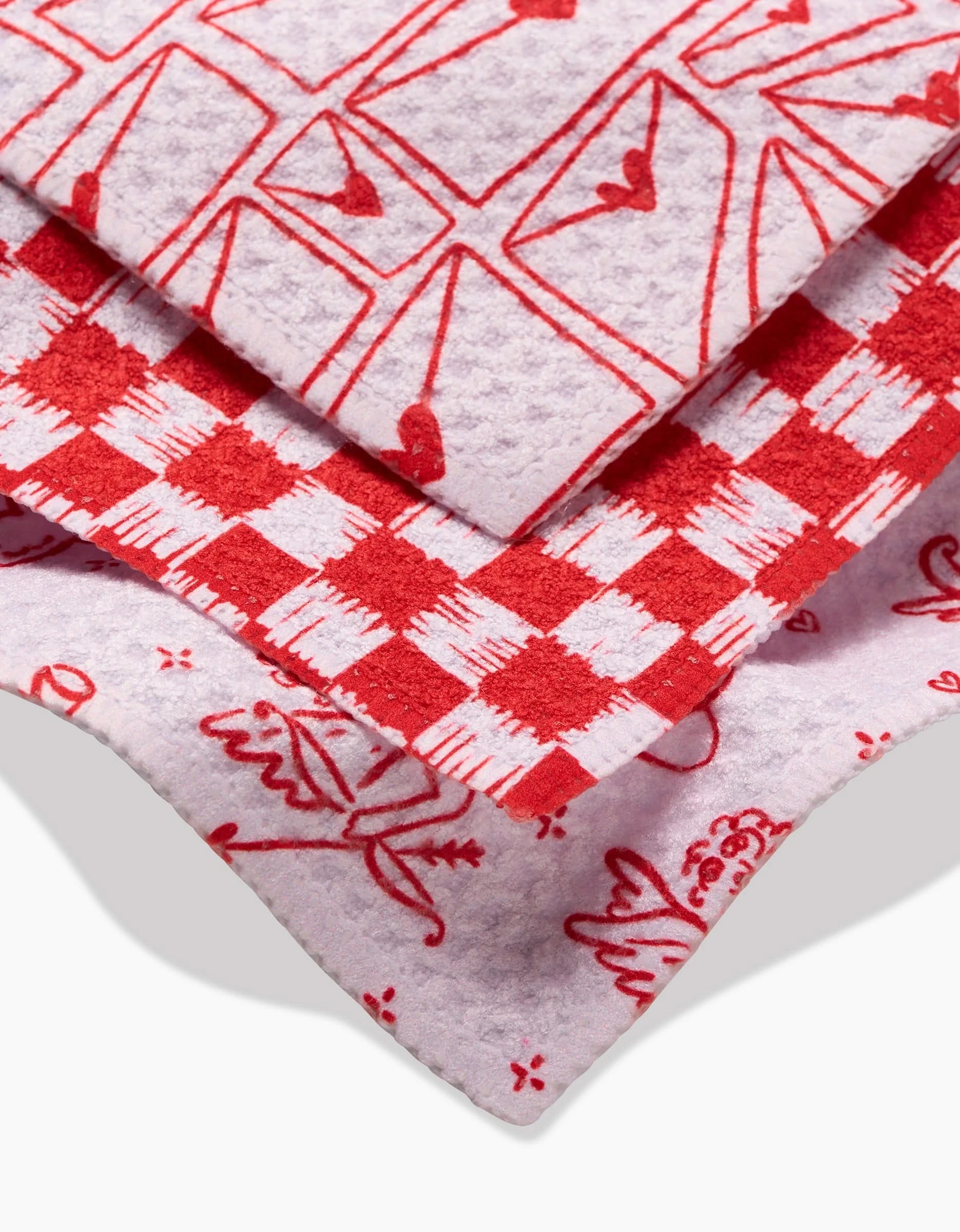 Kayla Ann’s Cupid’s My Valentine includes three stacked dishcloths made from recycled materials, each with unique red and white patterns—hearts and shapes on top, checkered middle, and whimsical flowers and objects on the bottom.
