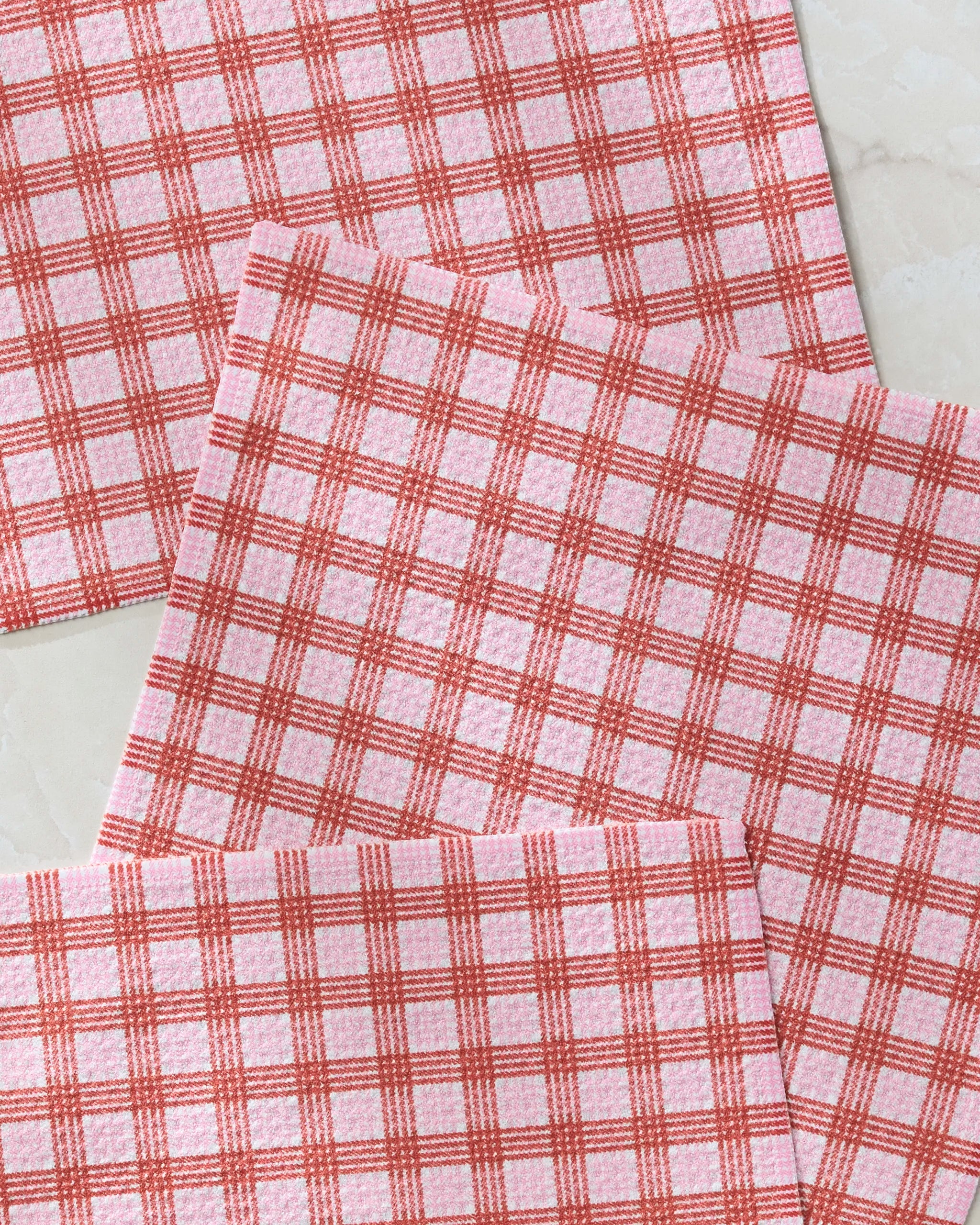 Geometry’s Cupid's Plaid napkins, made from recycled materials, feature a red and white checkered design and are arranged in an overlapping pattern on a light-colored surface.