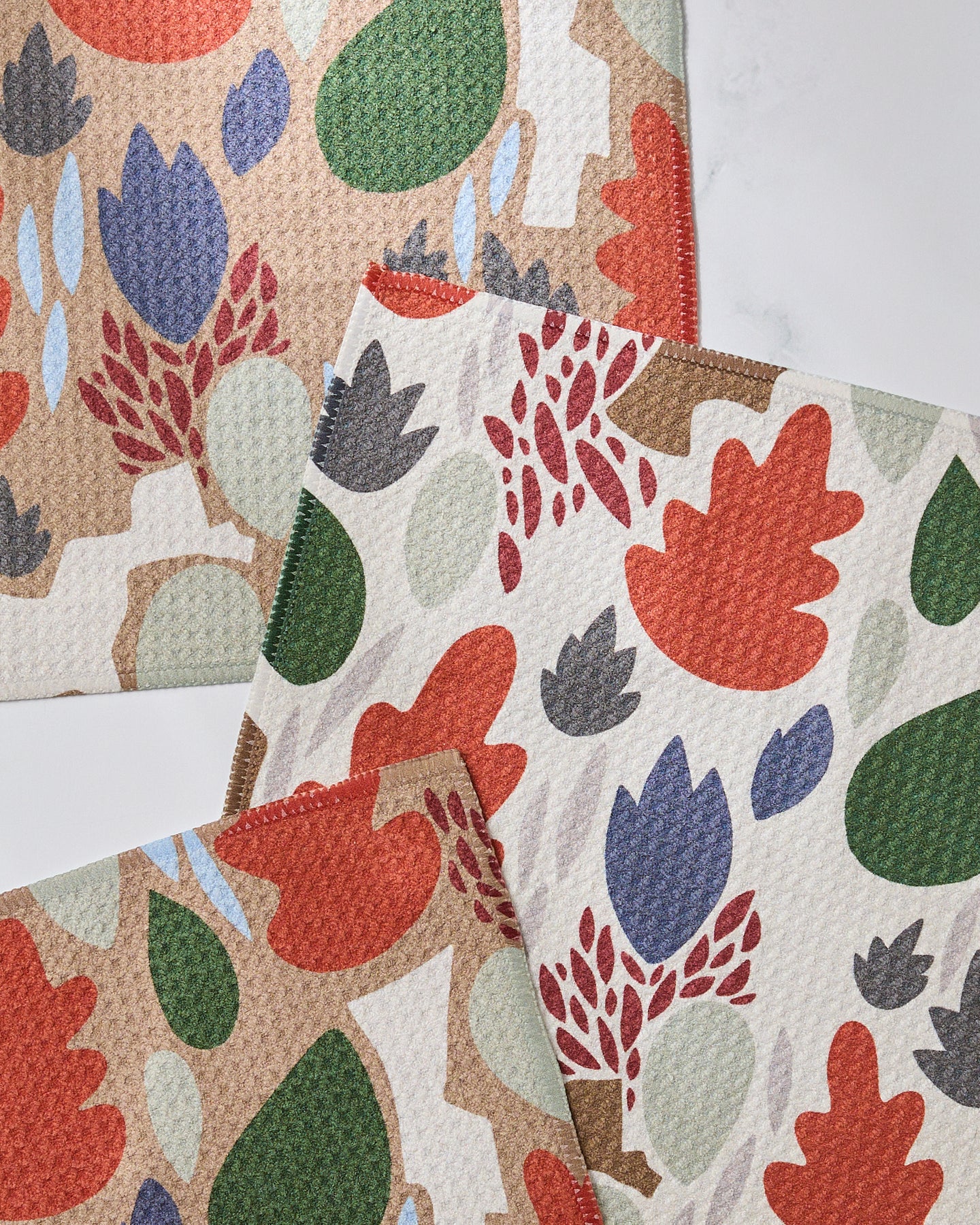 The Geometry Cut Paper Leaves set includes three super-absorbent, textured dishcloths featuring abstract botanical patterns in red, green, blue, and beige. Leaf and plant designs overlap on a light background.