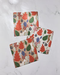 Three super-absorbent Geometry Cut Paper Leaves dishcloths, featuring colorful leaf and plant patterns in red, green, purple, and beige, are arranged on a white marble surface.