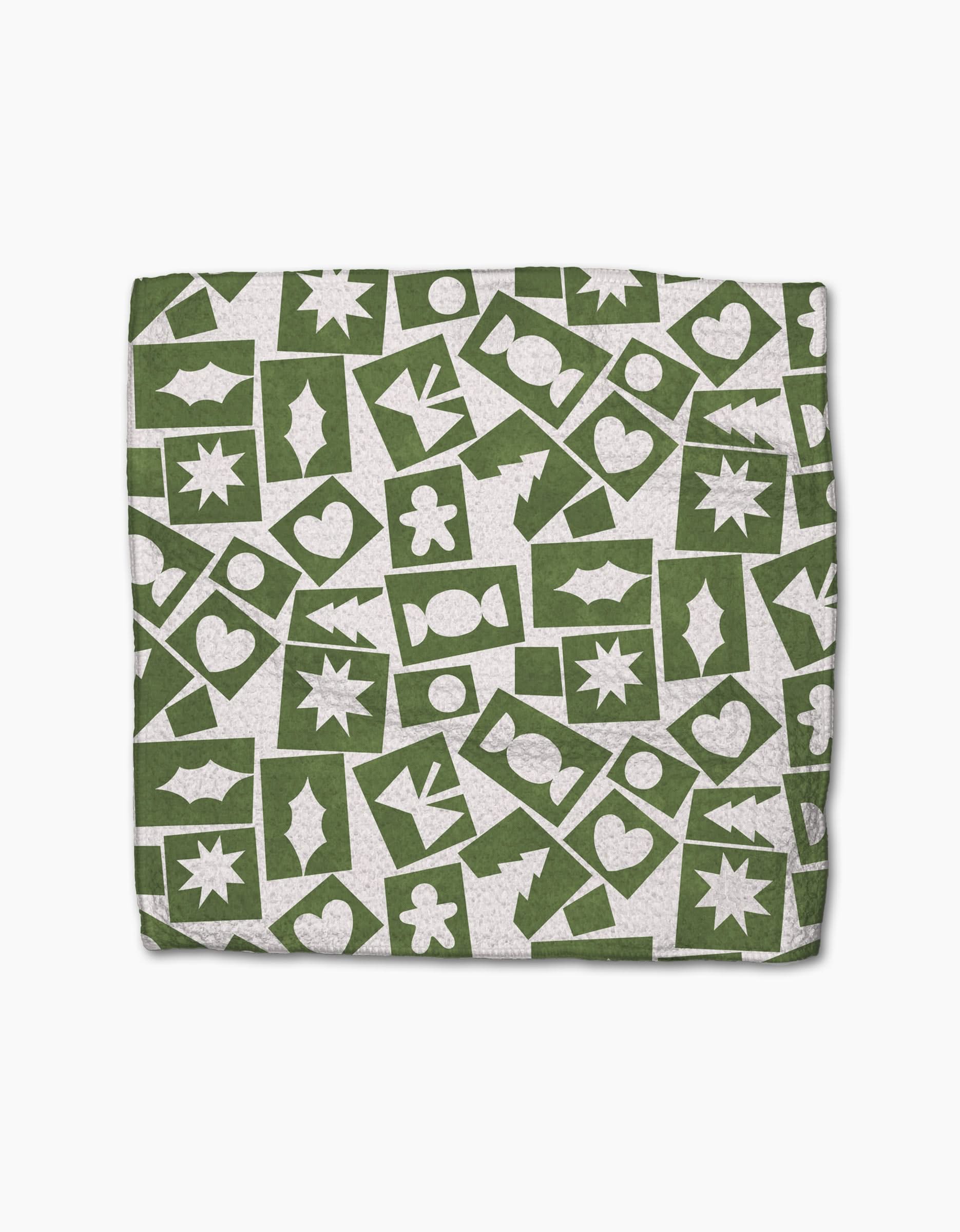 The Cutout Christmas napkin by Claire Iglesias features a green background with white festive shapes—stars, hearts, holly leaves, candy, and gingerbread figures—in a scattered pattern. Made from recycled materials.