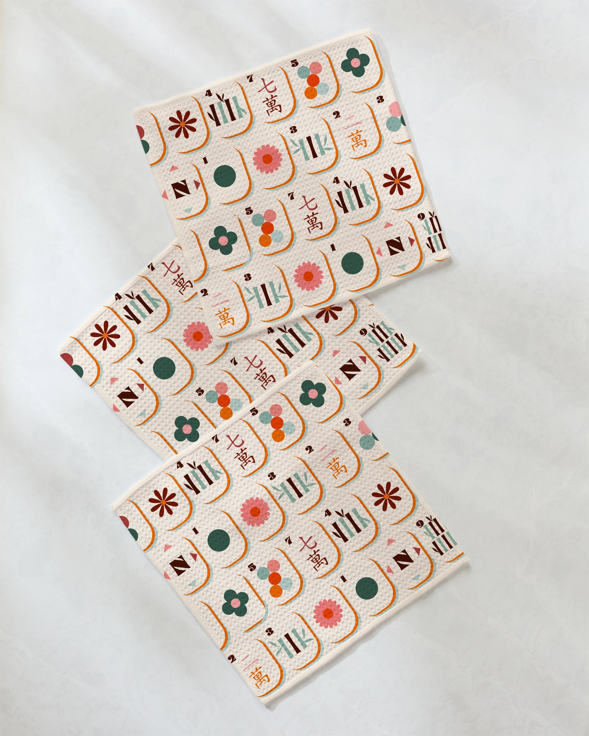 Three folded cloths from Geometry’s Cypress Tile Toss super-absorbent dishcloth set, each featuring a colorful pattern of flowers, circles, lines, and Japanese characters, are arranged diagonally on a light, textured surface.