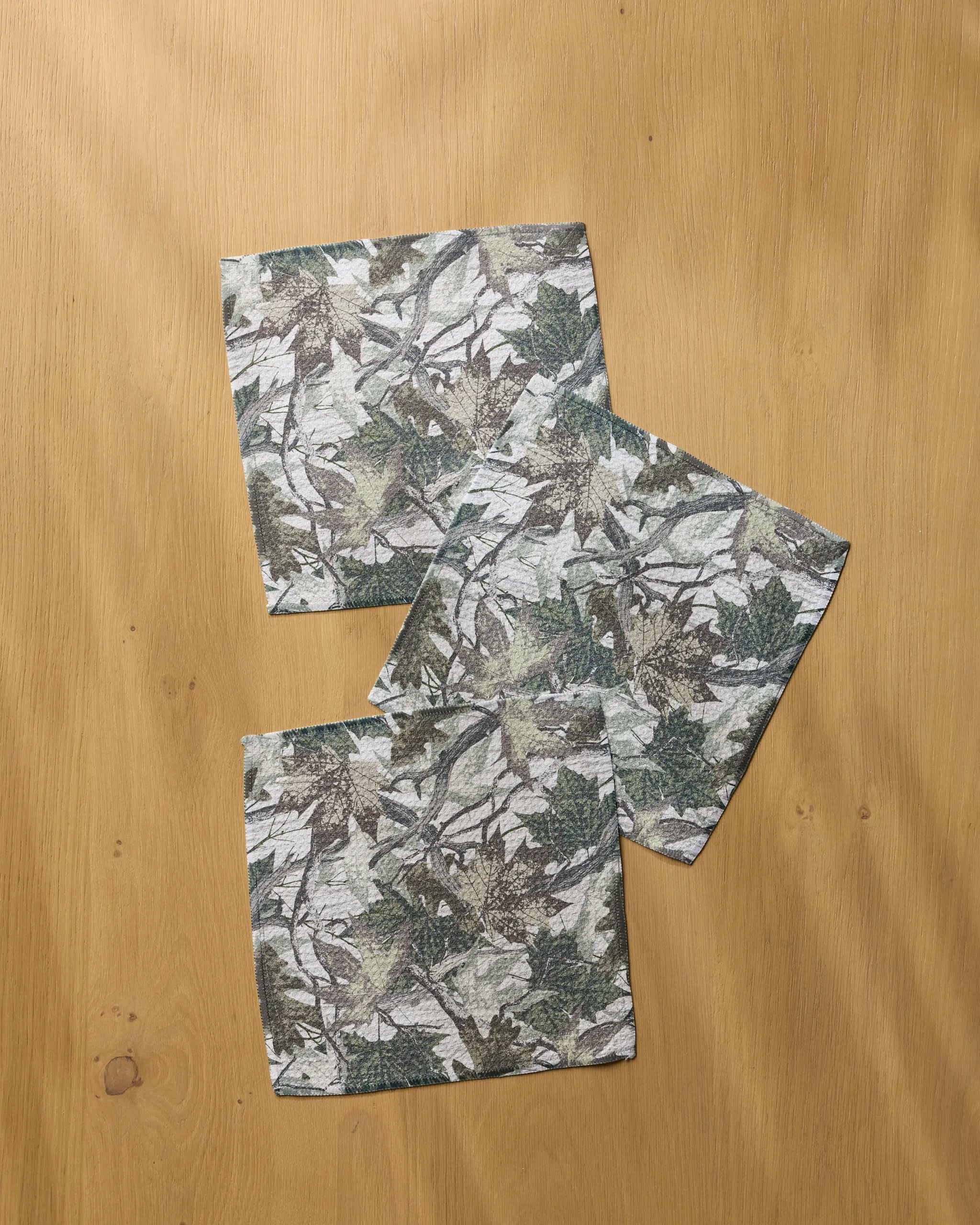 Three square napkins from Geometry’s "Dad Camo" set, featuring a green and gray leafy camouflage pattern, are displayed on a light wooden surface.