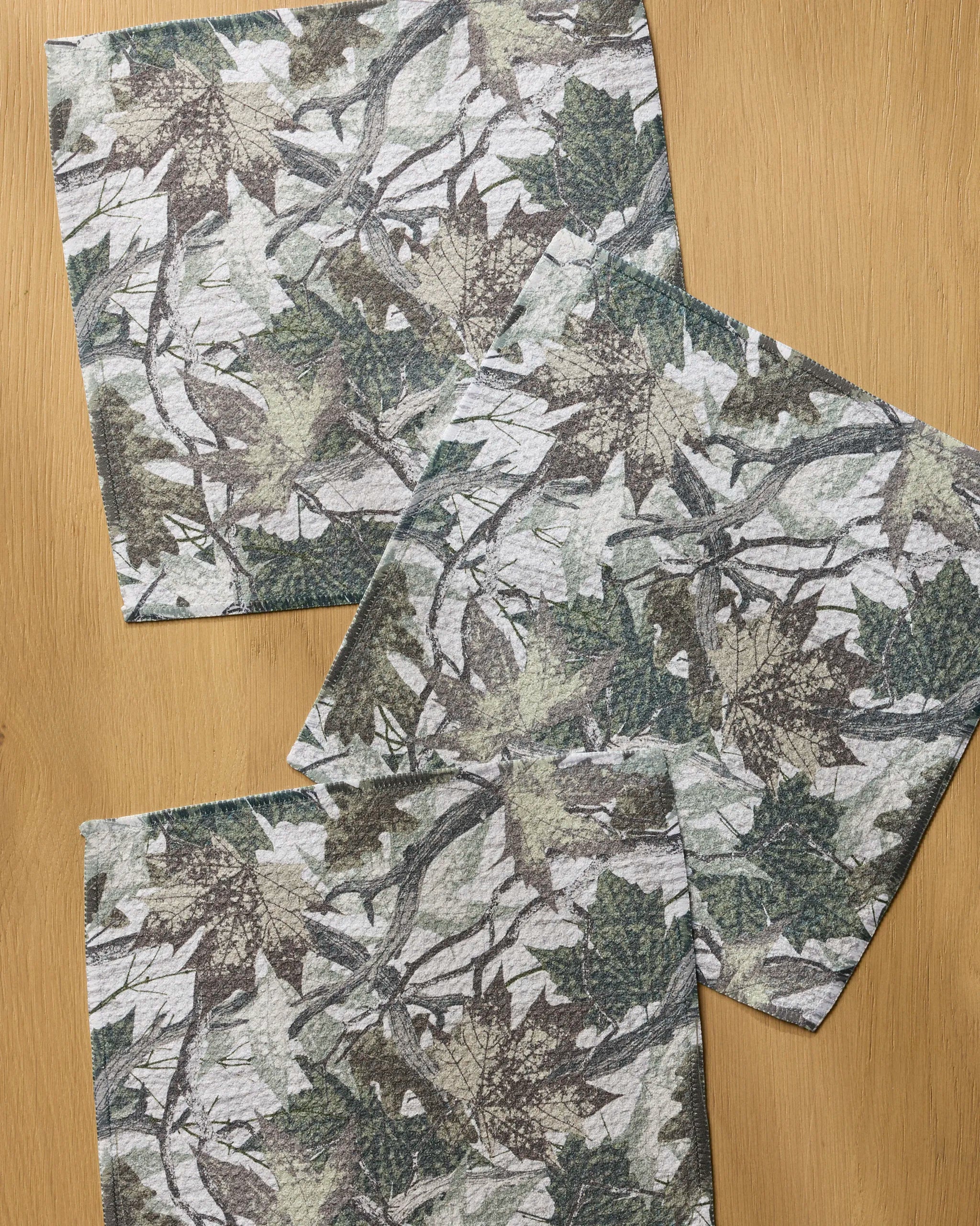 Three square Dad Camo towels from Geometry, featuring a green, brown, and gray camouflage leaf pattern, are arranged on a light wood surface—two overlapping diagonally and one upright—for a stylish, absorbent kitchen set.