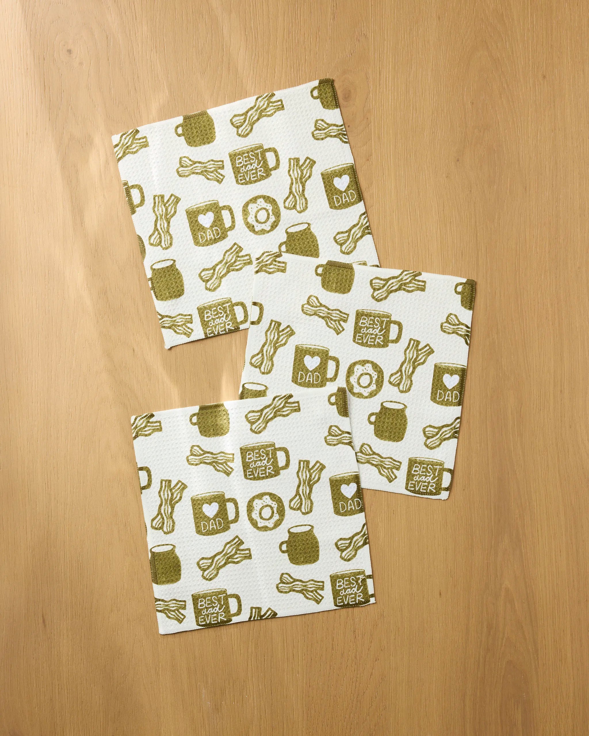 Three square napkins from Lisa Nelson’s Dad Fuel set, made of recycled dishcloth, show green mugs, bacon, donuts, and phrases like “Best Dad Ever” and “I ♥ Dad,” arranged on a light wood surface.
