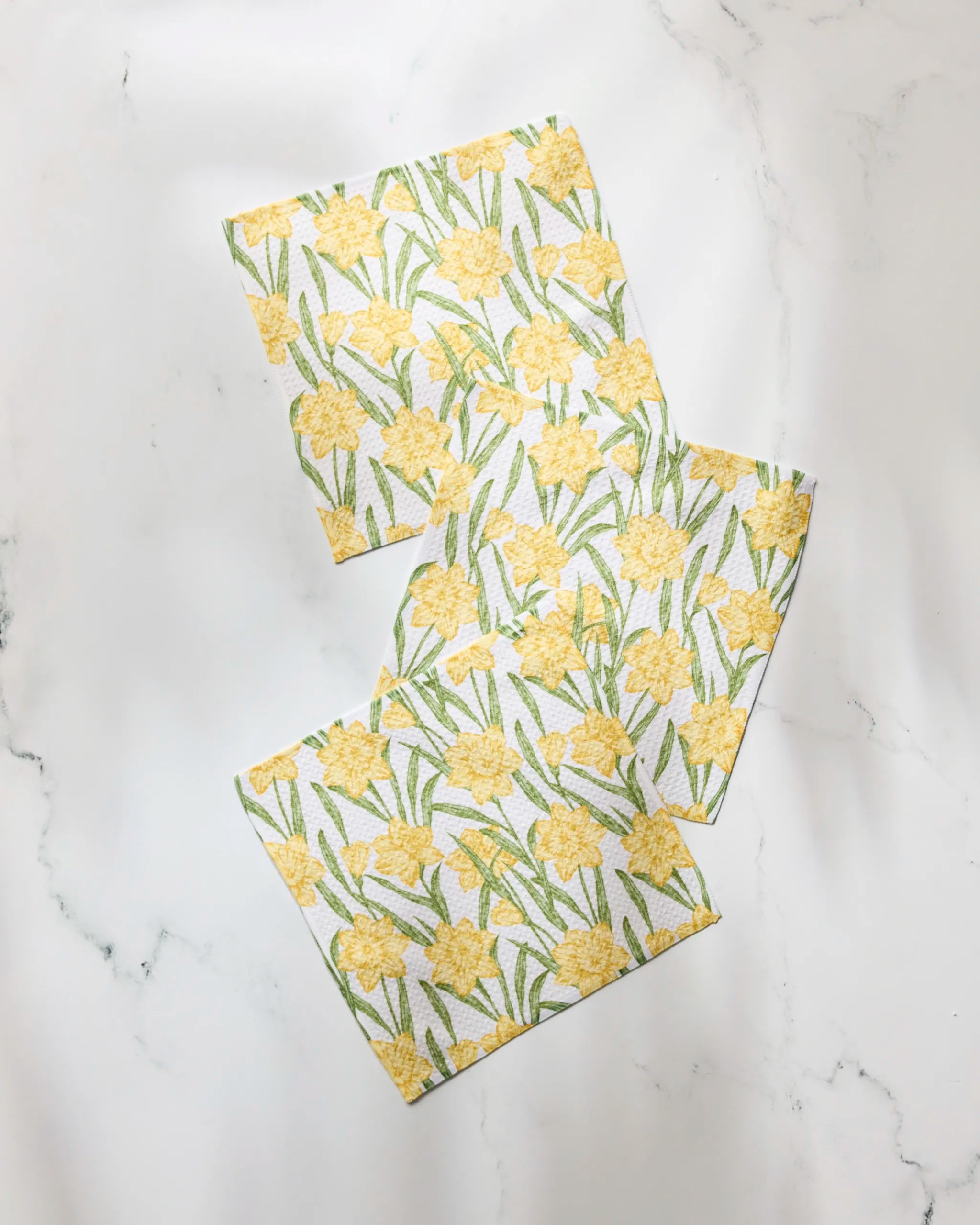 Three square Geometry Daffodil Darling towels, featuring yellow daffodils and green leaves, are arranged diagonally on white marble. This super-absorbent set is crafted from recycled materials for stylish, sustainable kitchen use.