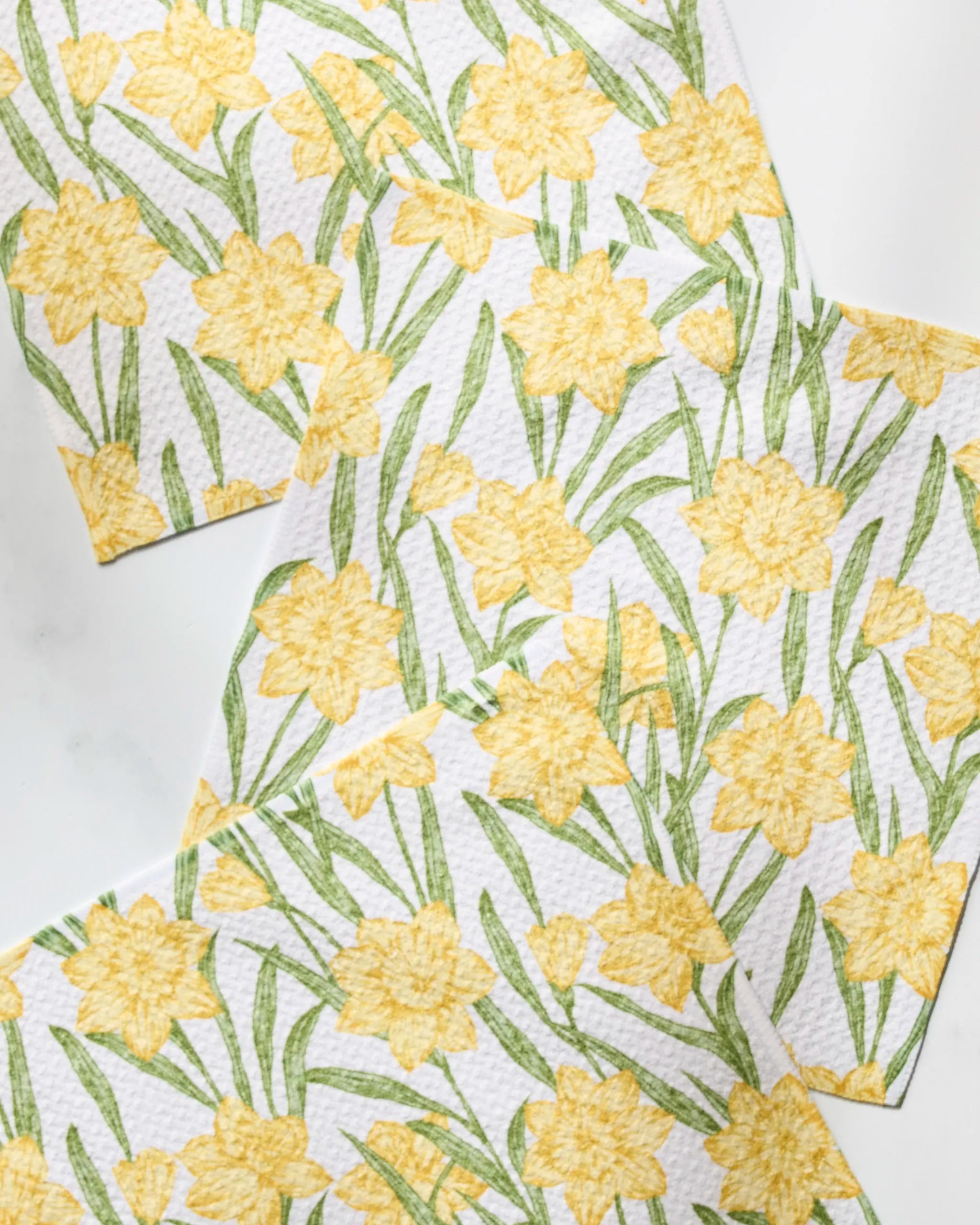 Geometry’s Daffodil Darling set includes four recycled paper napkins, each featuring yellow daffodils and green leaves in a staggered, overlapping design on a white background.