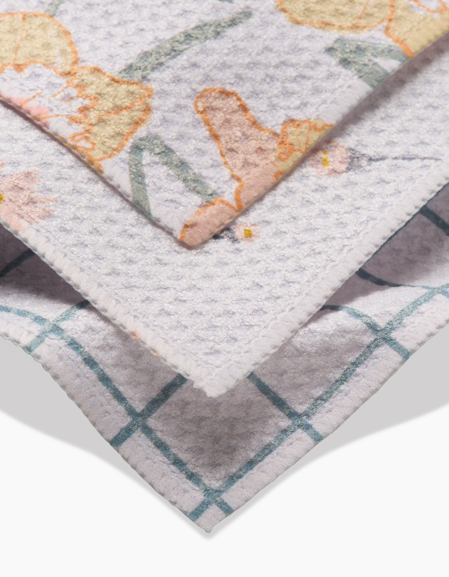 Close-up of three stacked Hufton Studio Daisy Dust dishcloths: a floral pattern in soft orange and green, a plain light grey, and a teal grid on white Geoweave fabric for super-absorbent cleaning.