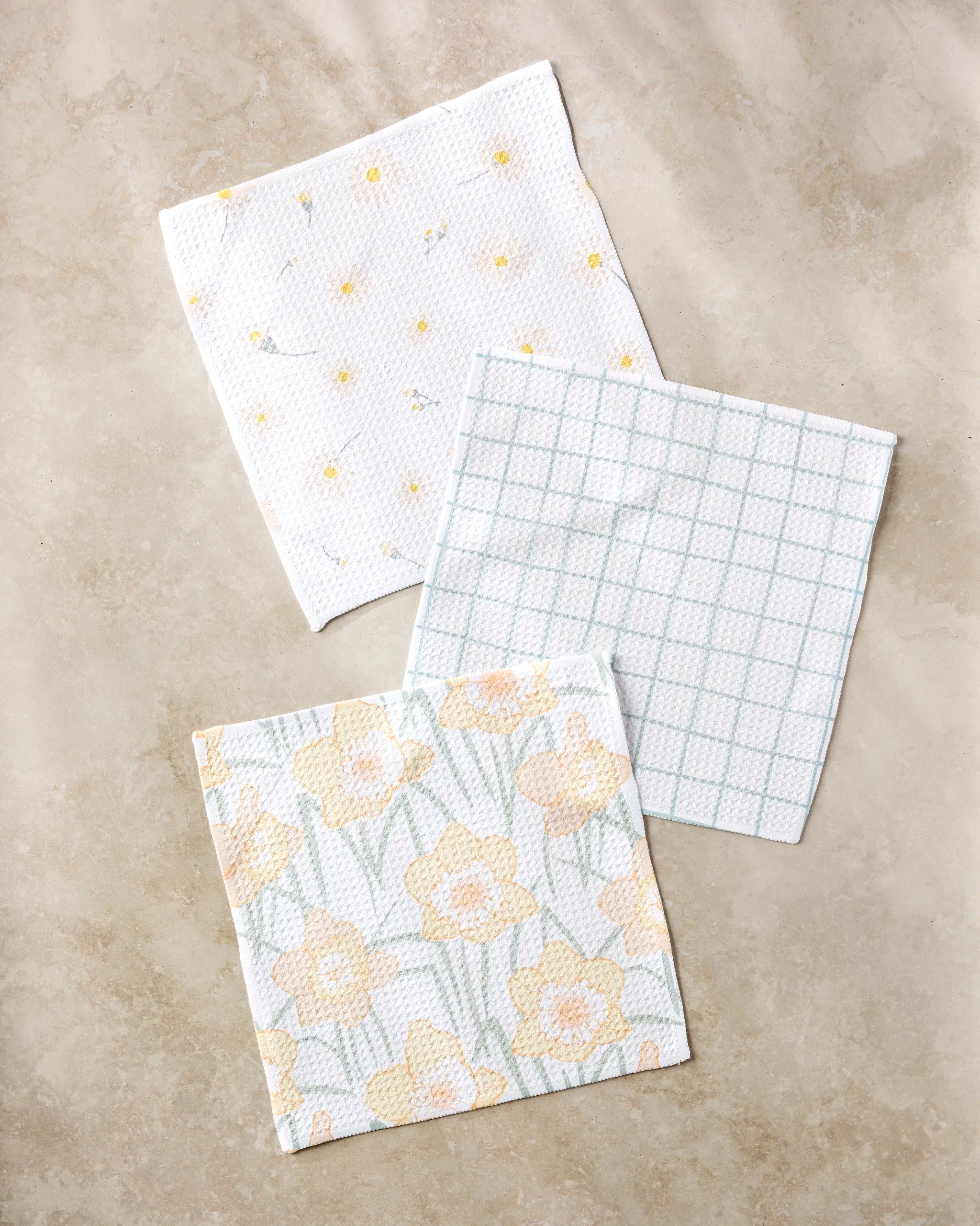 Three "Daisy Dust" patterned cloth napkins by Hufton Studio, made from recycled materials, are arranged on a beige surface—one with small yellow flowers, one in a blue grid pattern, and one with large yellow daffodils.