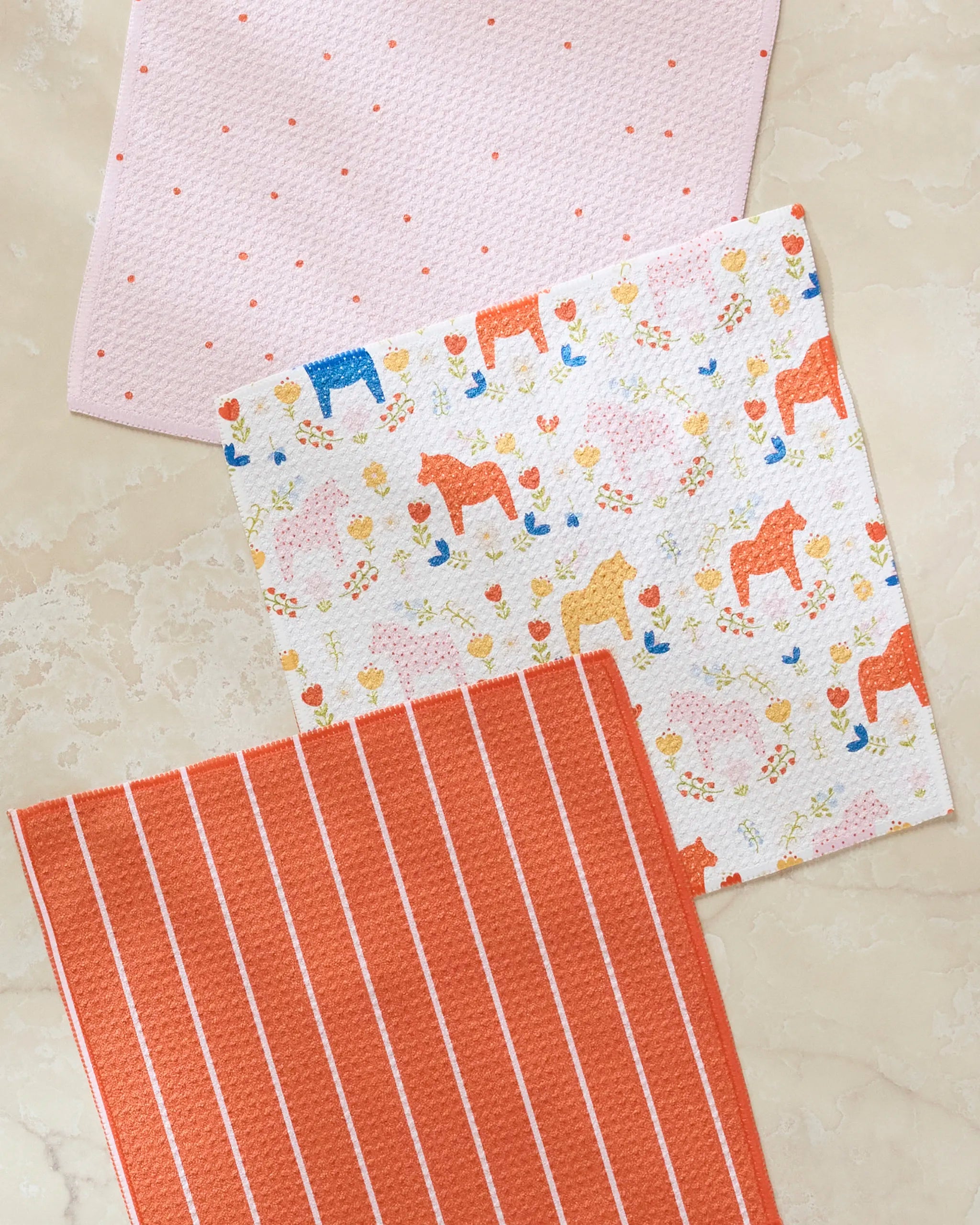 The Sara Mari Dala Garden Floral set includes three super-absorbent dishcloths—pink with dots, white with horses and flowers, and orange with stripes—all made from recycled materials.