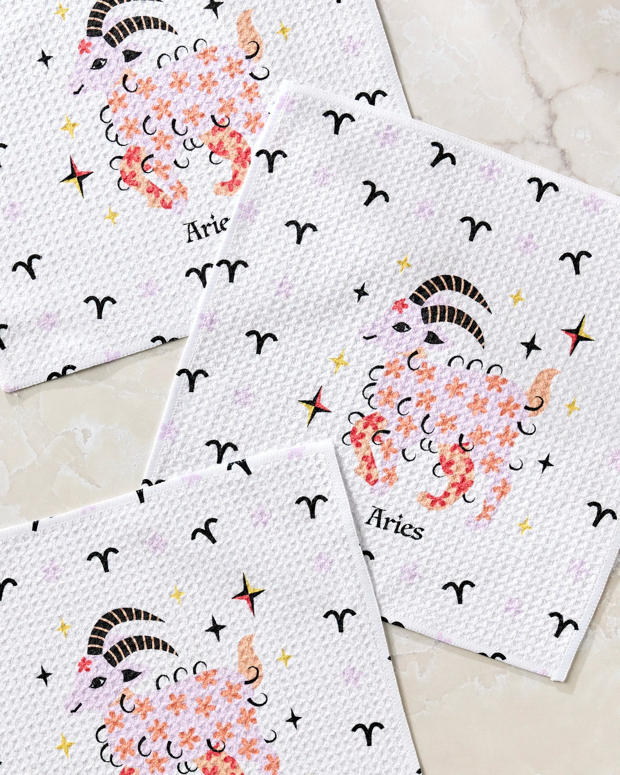 Heather Zhou’s "Daring Aries" set features three white recycled dishcloths with floral sheep and curled horns, Aries zodiac symbols, and colorful stars on a light marble background.