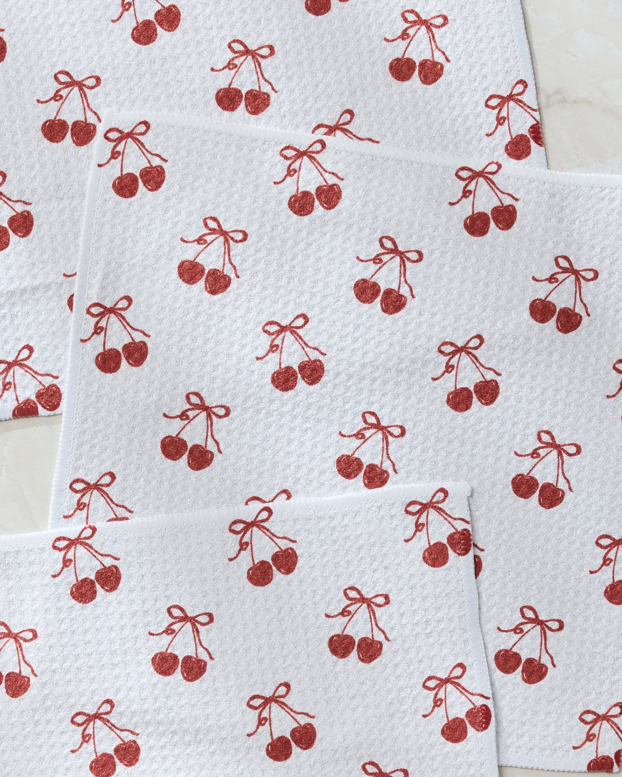 Geometry’s “Darling” white paper napkins feature a repeating red cherry pattern with each pair tied by a bow, overlapping on a light surface—an ideal match for any dishcloth set.