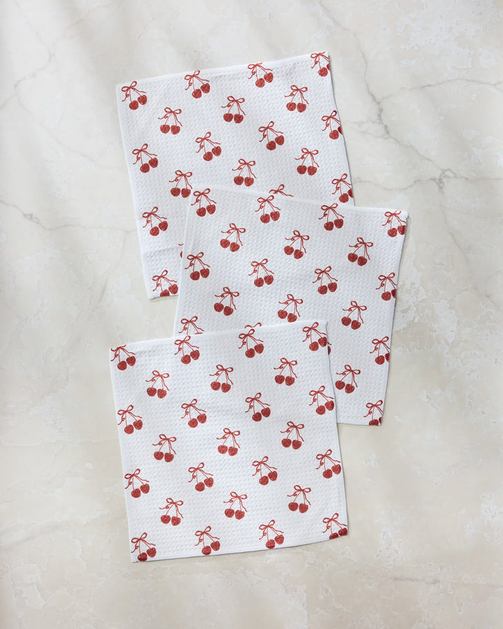 Three "Darling" napkins by Geometry, made from recycled materials and featuring a repeating pattern of red cherries and small bows, are arranged on a light marble surface.