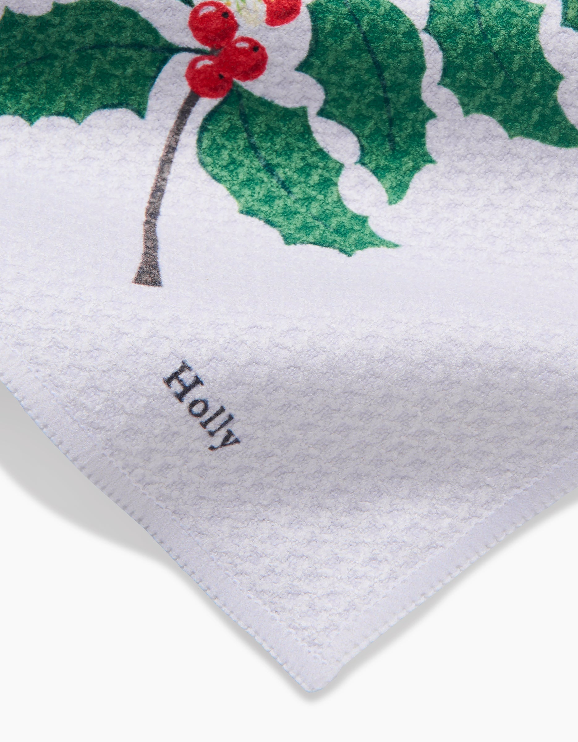 The December Holly dishcloth by Linda Fox is made of white Geoweave fabric and features a printed design with green holly leaves, red berries, and Holly in black on one corner.