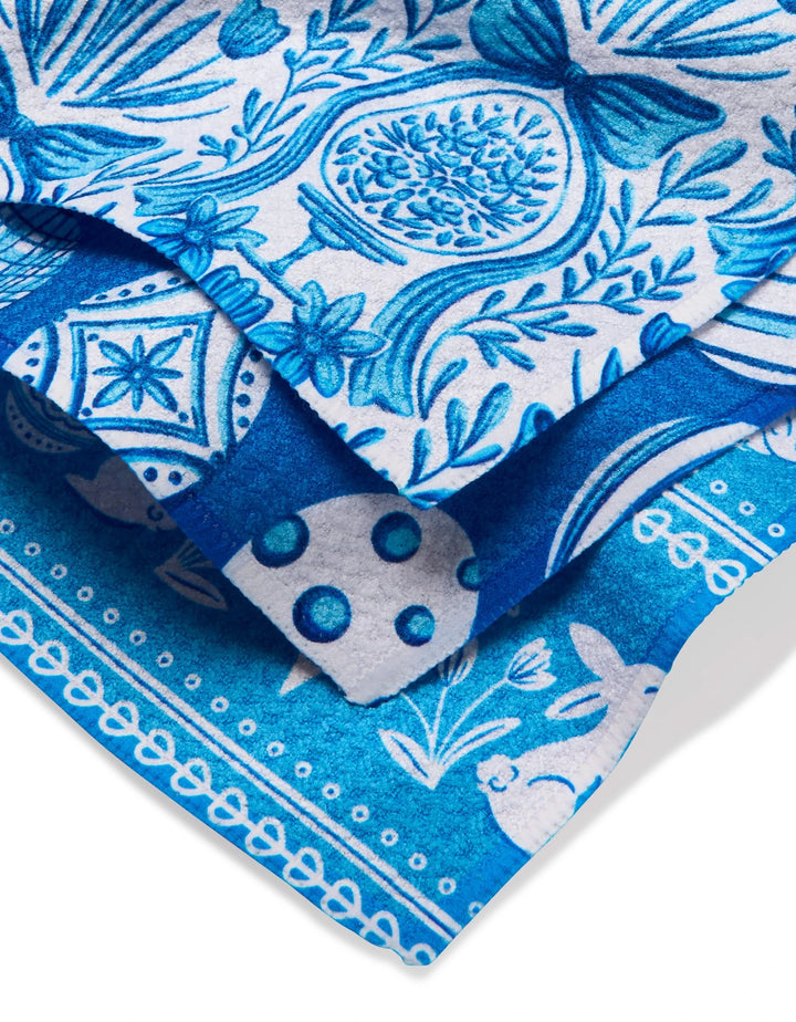The Delft Blue Easter Eggs and Bunnies dishcloth set by Sarah Daugherty features three folded towels with blue and white patterns of flowers, birds, hearts, and geometric shapes—crafted from recycled materials for an eco-friendly touch.