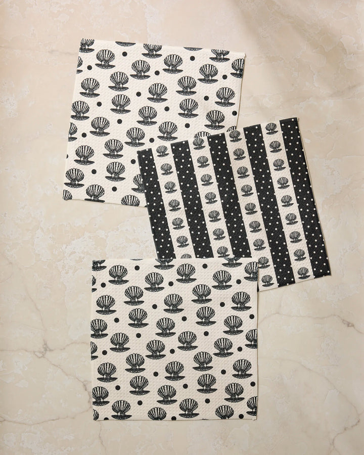 Four Disco Pearls cloth napkins by Geometry, made from recycled materials, are set on a beige marble surface: two with black and white seashell patterns and two with black and white stripes and dots.