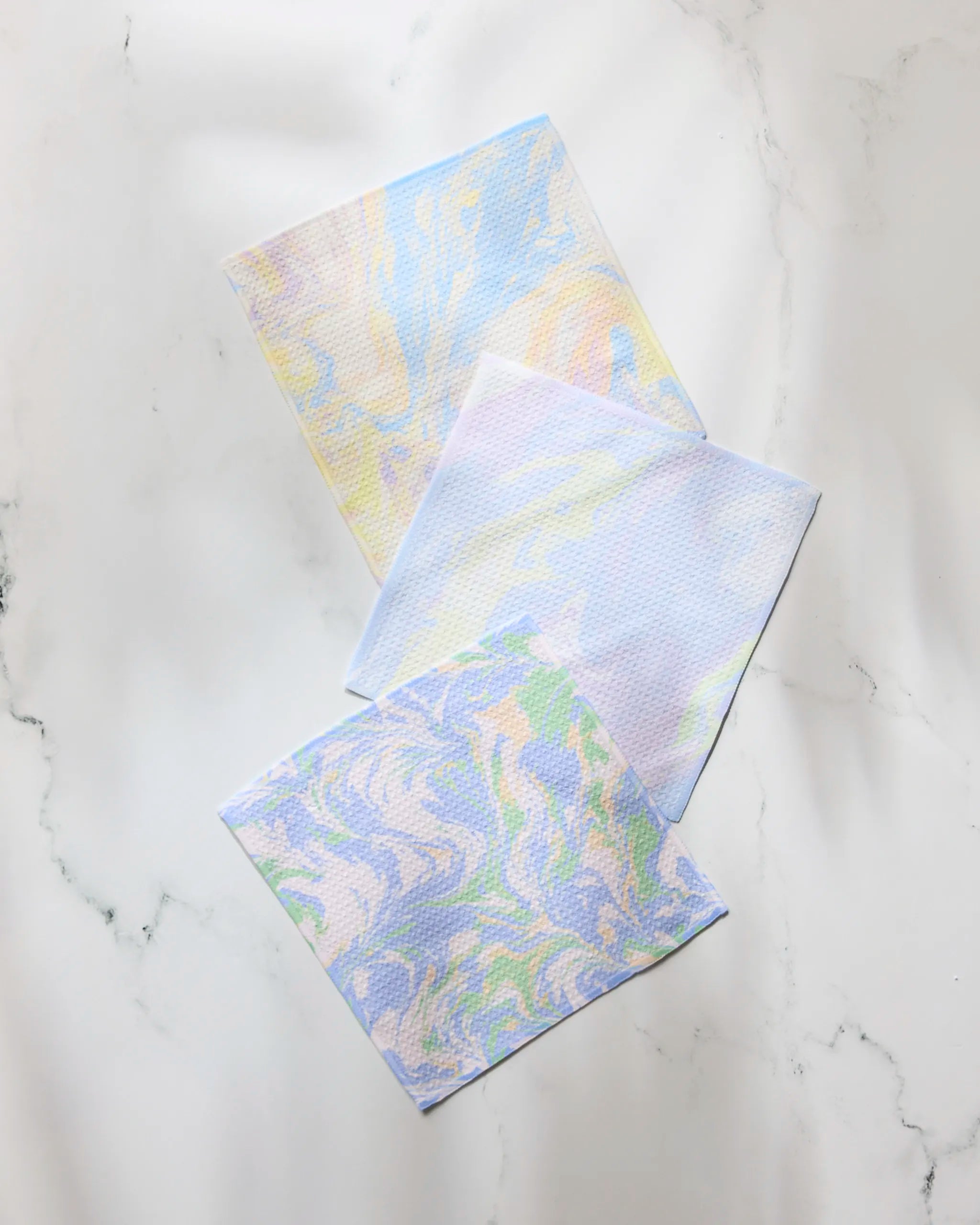 The Geometry Dream Skies set features three rectangular dishcloths with pastel marbled patterns in blue, purple, yellow, and green tones—stylishly crafted from recycled materials and displayed on a white marble surface.