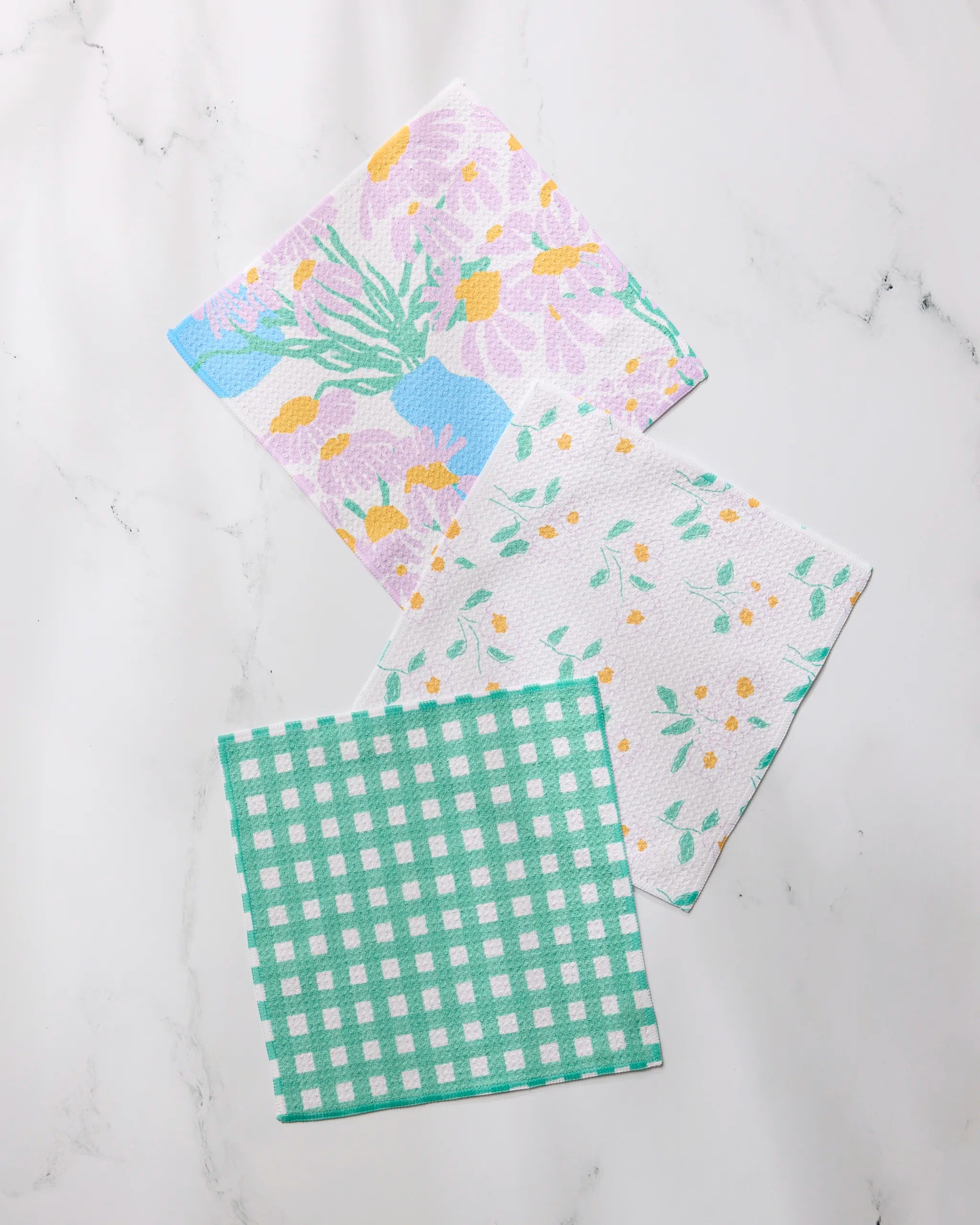 Three "Dreaming of Florals" napkins by Geometry—featuring a floral vase print, delicate yellow and green blossoms, and a green check pattern—are laid out on white marble. All are crafted from recycled materials.