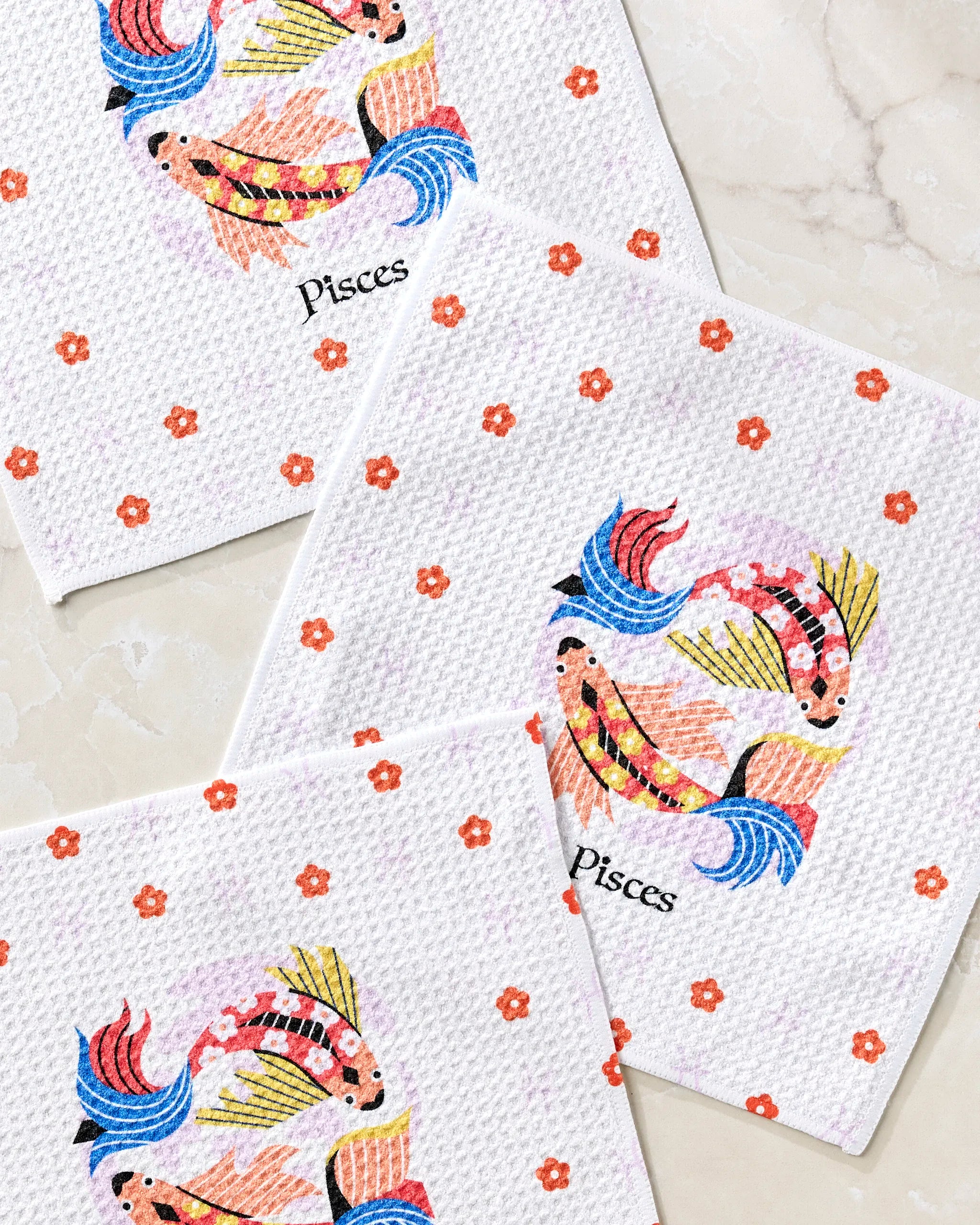 The “Dreamy Pisces” dishcloth set by Heather Zhou includes three white textured towels with orange flowers and vibrant fish in a Pisces-inspired circle, crafted from recycled materials and displayed on light marble.