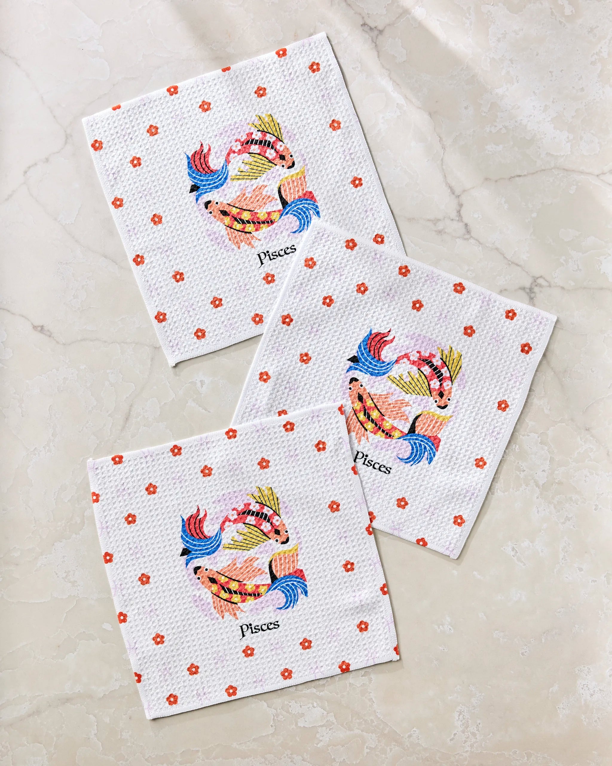 Three "Dreamy Pisces" white cloth napkins by Heather Zhou, made from recycled materials, feature a colorful Pisces fish design with small red flowers, arranged on a light marble surface.