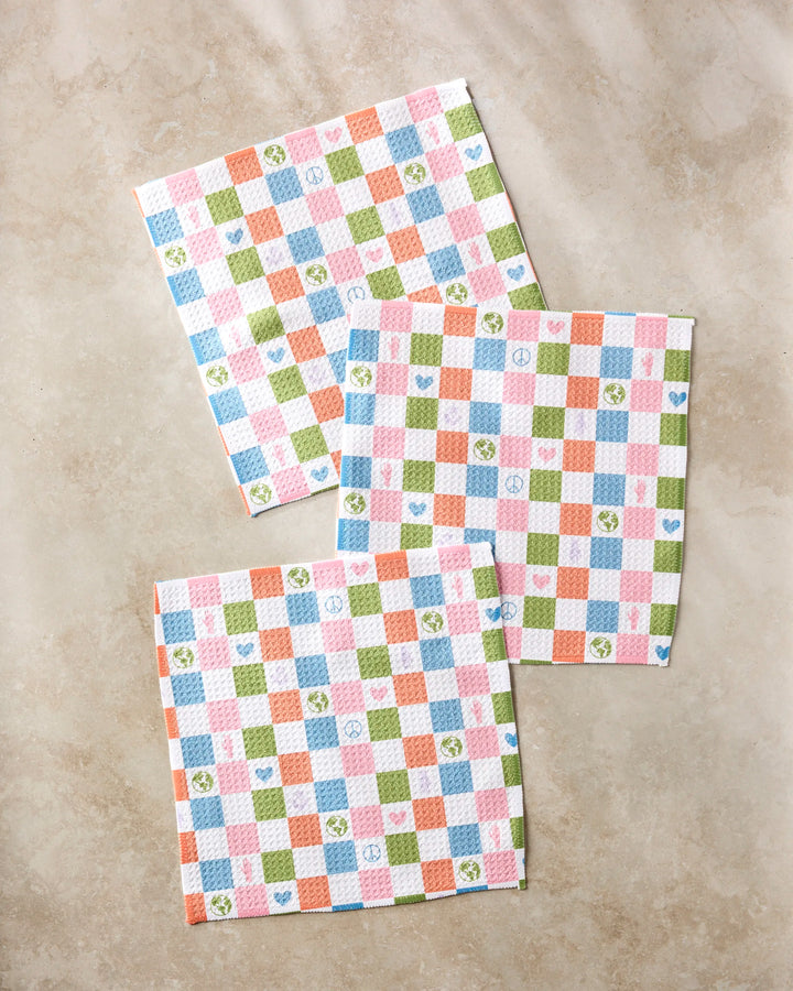 Three square napkins from Kayla Ann's Earth Day Checkers set, with vibrant checks and icons like hearts, stars, and flowers, are displayed on a beige surface—ideal as kitchen towels or recycled dishcloths.