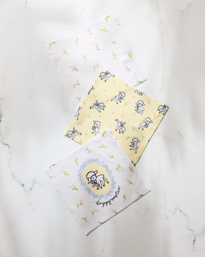 Three square Geometry dishcloths from the Easter Lamb collection are displayed on white marble: two with floral prints, one yellow with sheep illustrations, and another featuring an embroidered sheep and "happy as a lark.