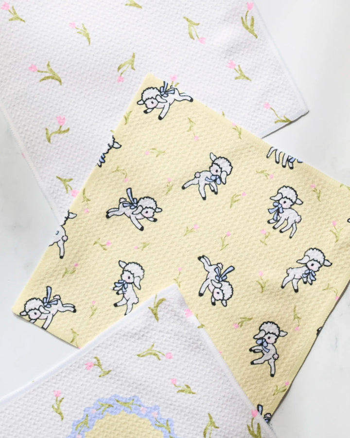 The Geometry Easter Lamb absorbent dishcloth set includes three textured cloths: two with a white background and green branches with pink buds, and one yellow cloth featuring white lambs with blue bows.