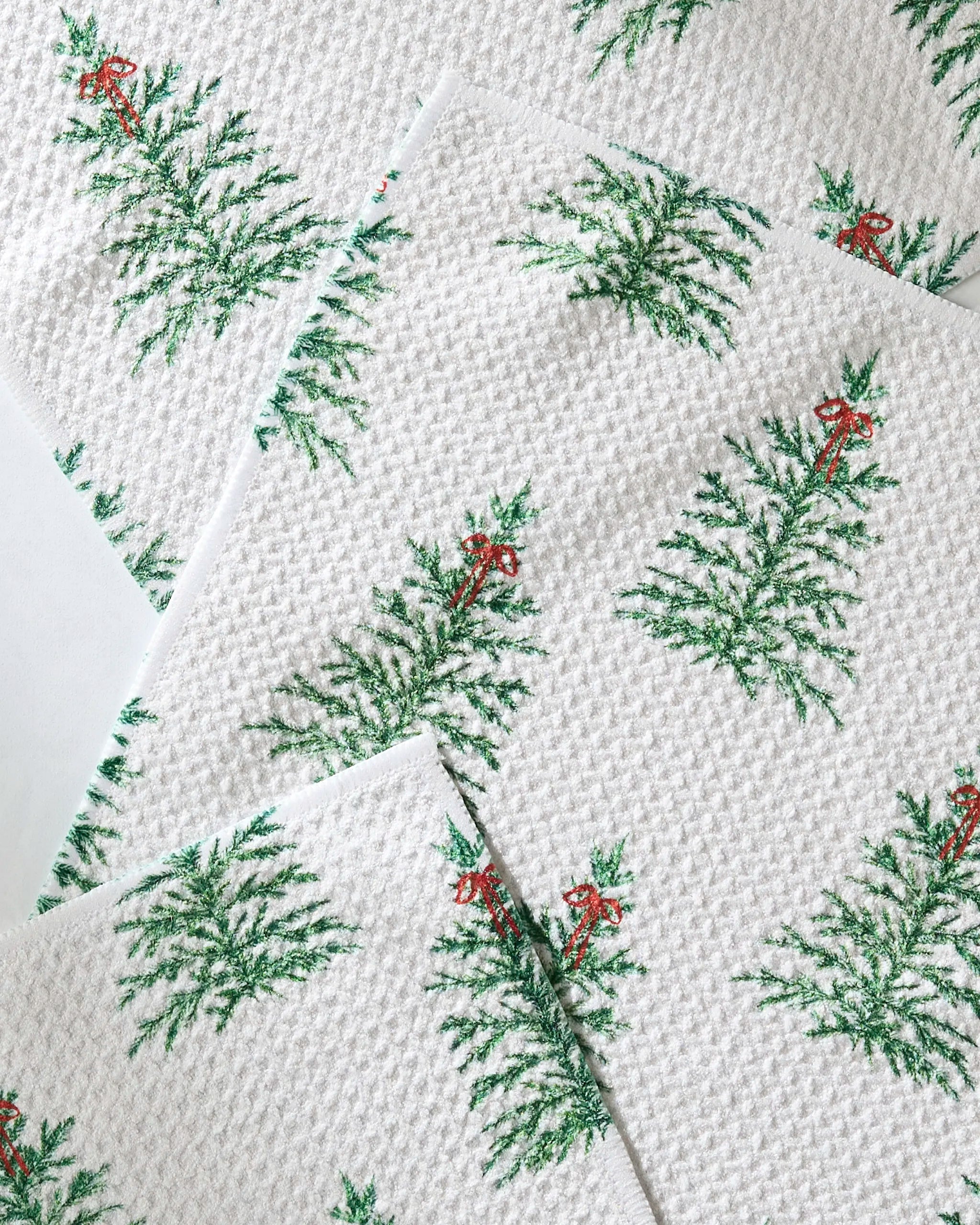 The Geometry Ellsworth napkins feature white textured fabric with green Christmas trees topped by red bows, overlapping at various angles for a festive touch to your holiday table.