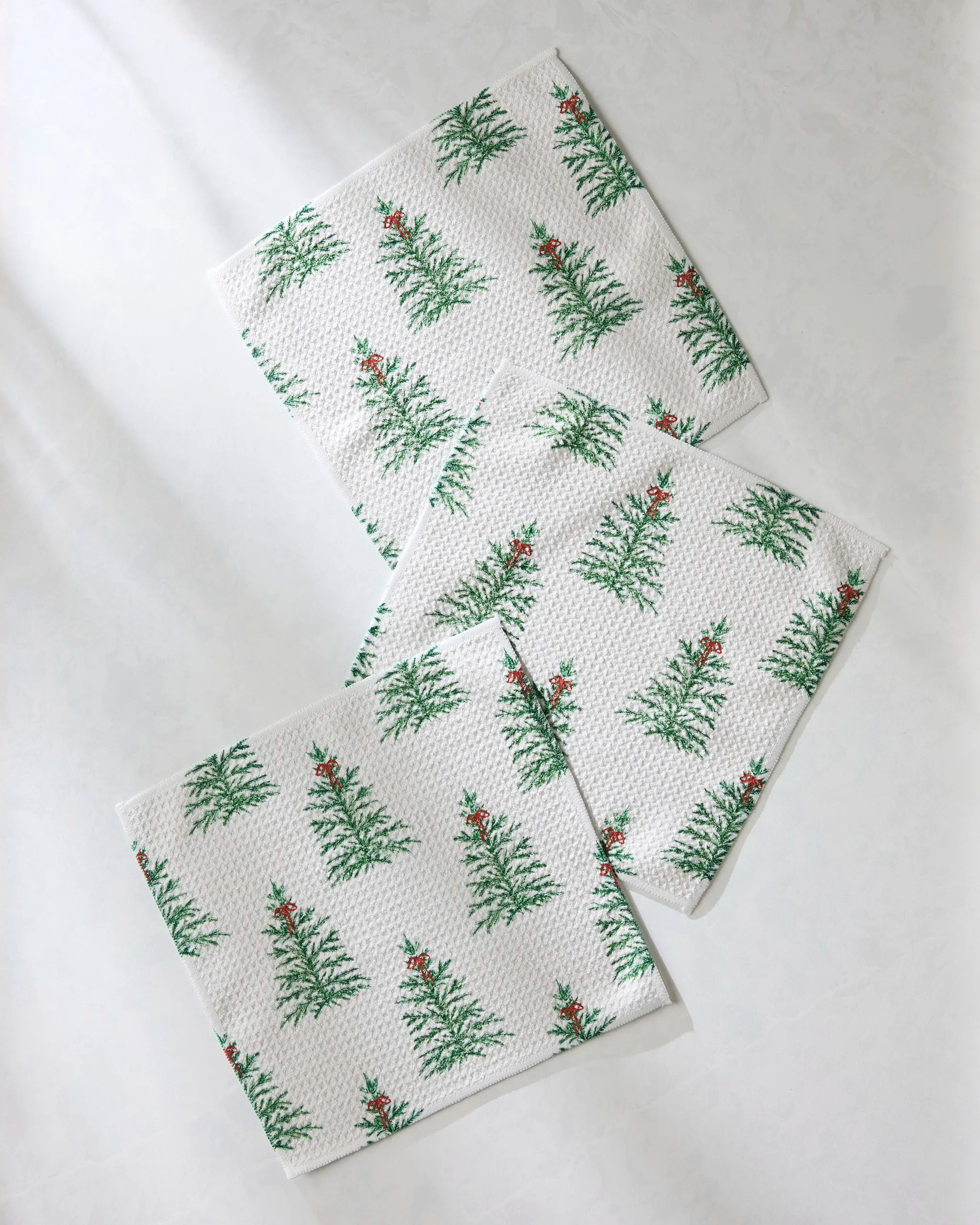 Three Ellsworth paper napkins by Geometry, featuring green Christmas trees and red berries, are arranged with a slight overlap on a light surface—ideal for pairing with your favorite dishcloths.