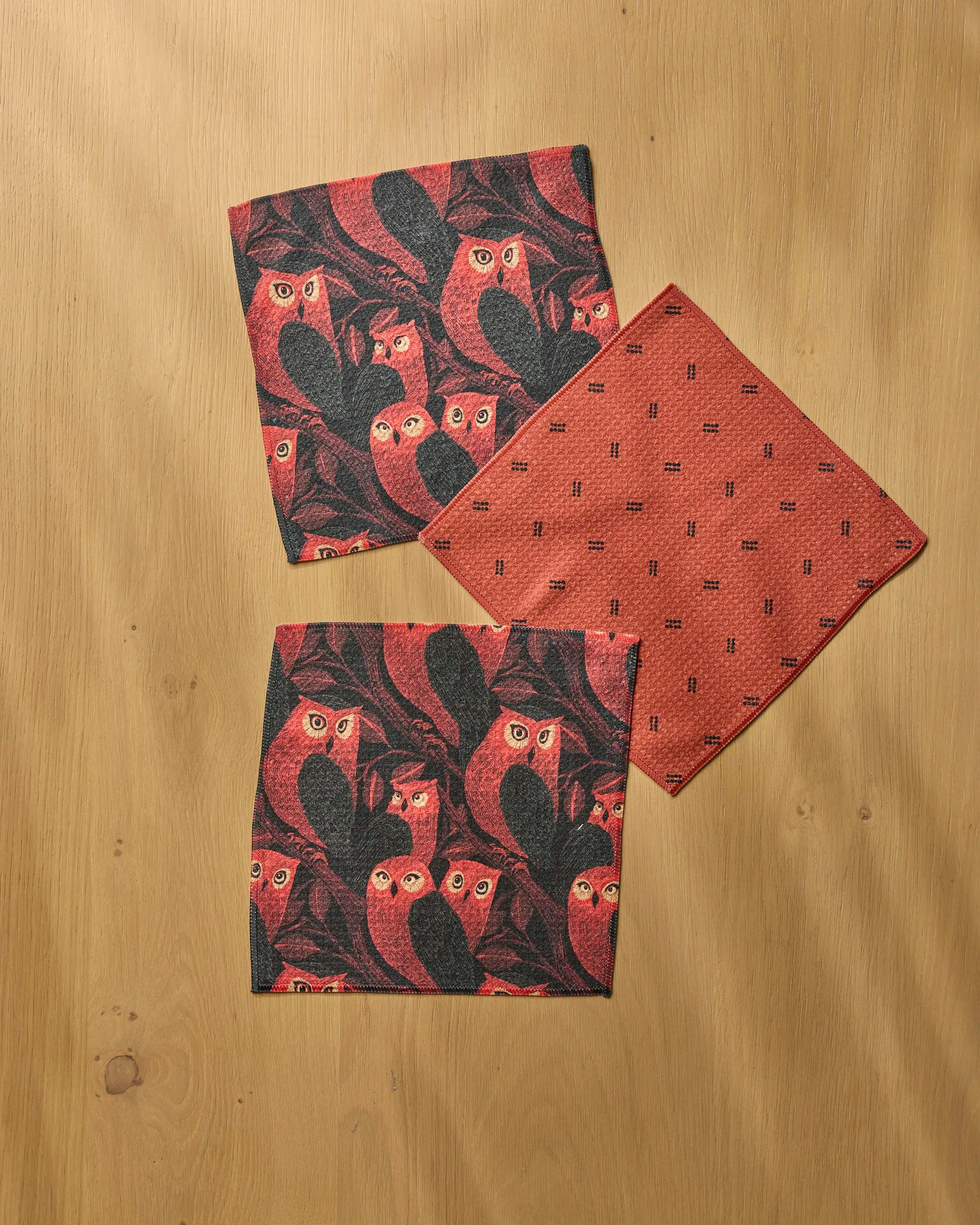 Three folded Geometry Ember Owl napkins on wood: two feature a red and black owl pattern, while one has a subtle red geometric design, all made from recycled materials for eco-friendly style.
