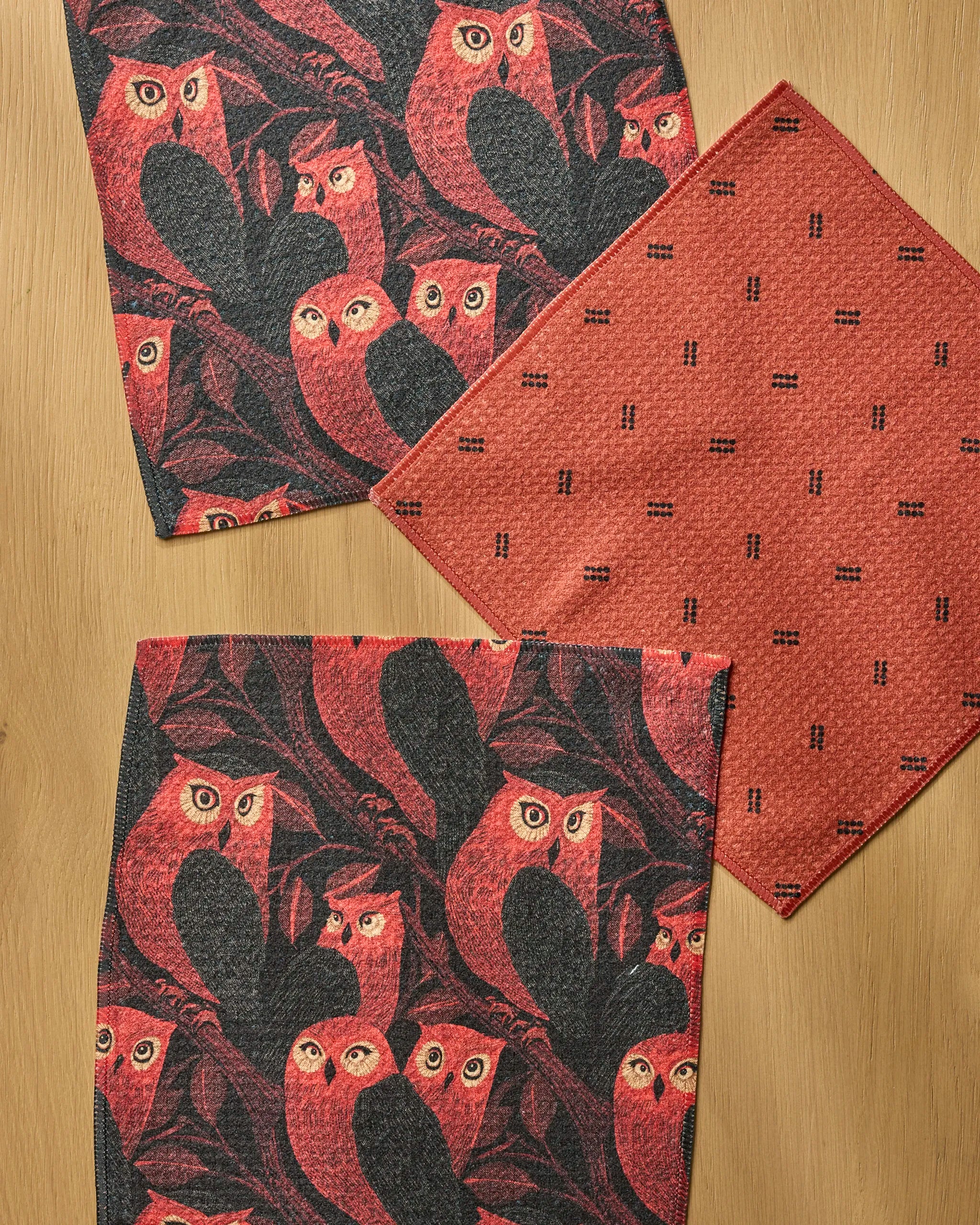 The Geometry Ember Owl set features three eco-friendly, fast-drying dishcloths: two with a red and black owl pattern and one plain red with a small black design, all made from recycled materials.