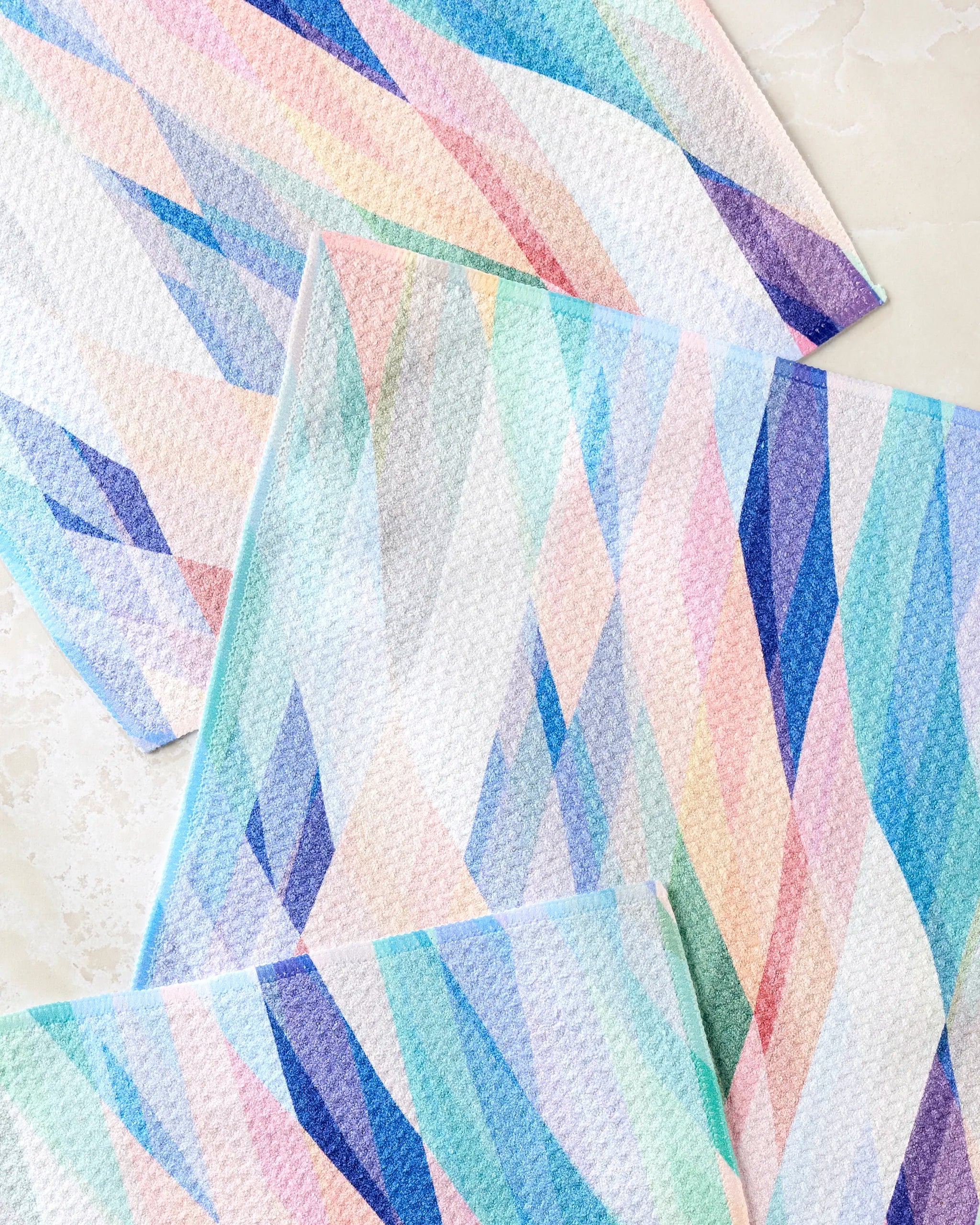Three Rachel Parker "Eventide" dishcloths with pastel geometric patterns in blue, pink, purple, and green are arranged on a light surface.