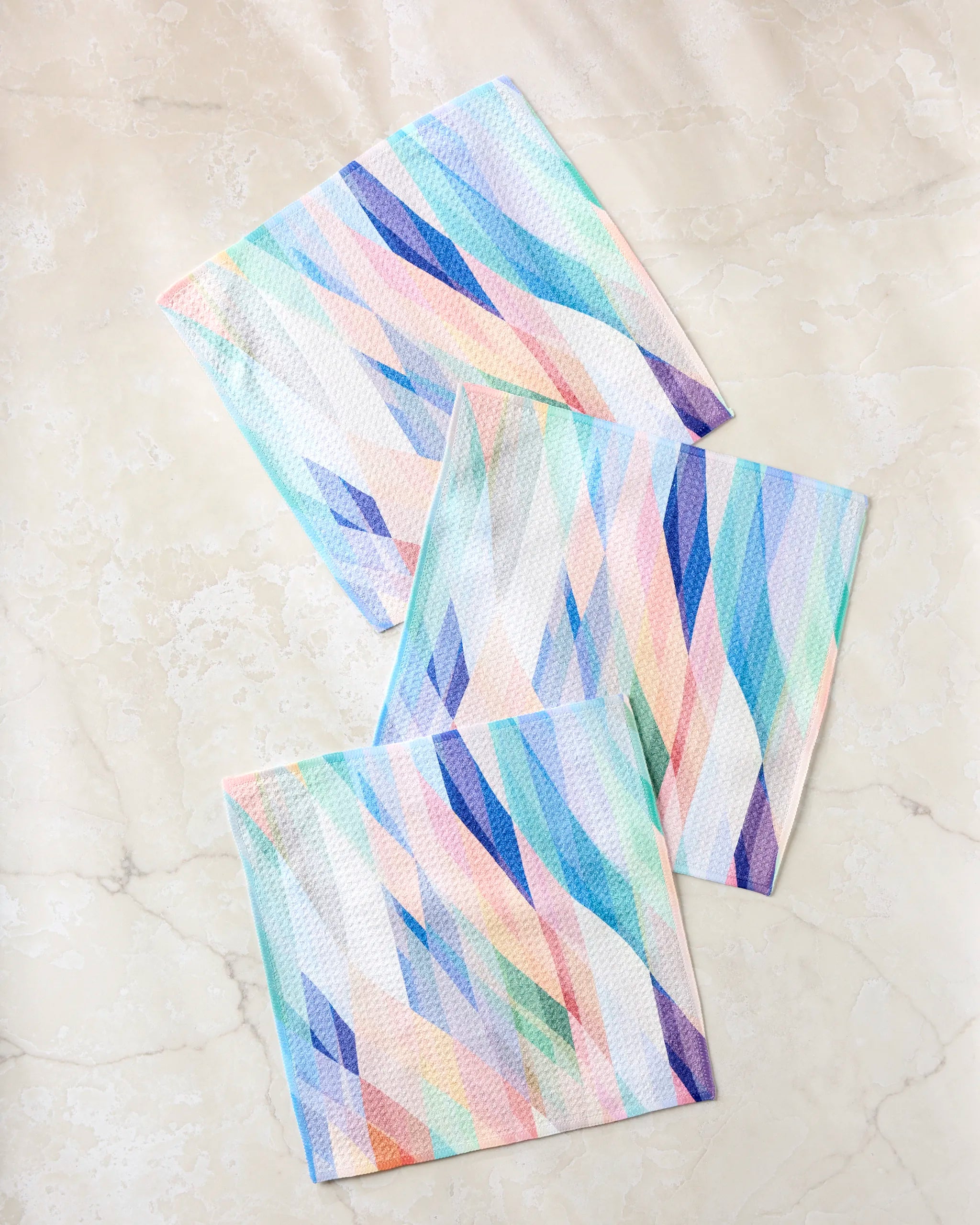 Three Eventide dishcloths by Rachel Parker, each textured and absorbent, feature pastel wavy and geometric designs in blue, pink, green, and purple on a light marble surface.