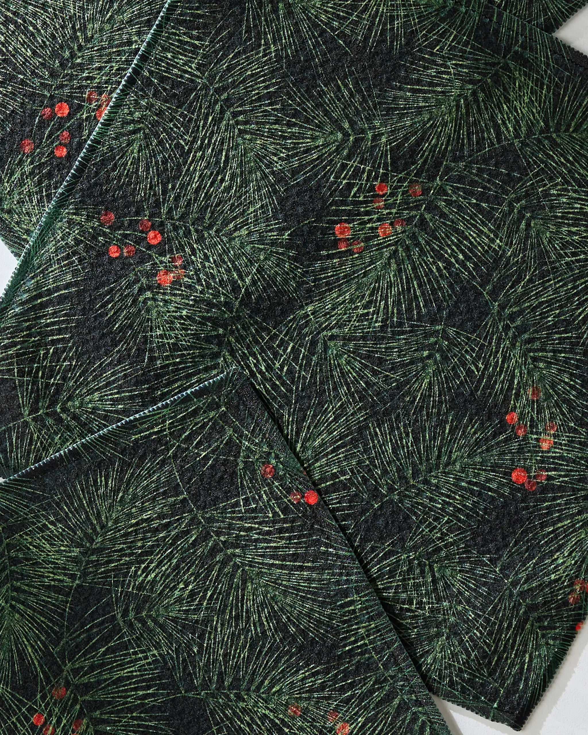 Close-up of Geometry’s Evergreen Pine dish towels with a green pine needle pattern and red berry clusters, arranged in overlapping layers. The texture and details of this absorbent dishcloth set stand out.