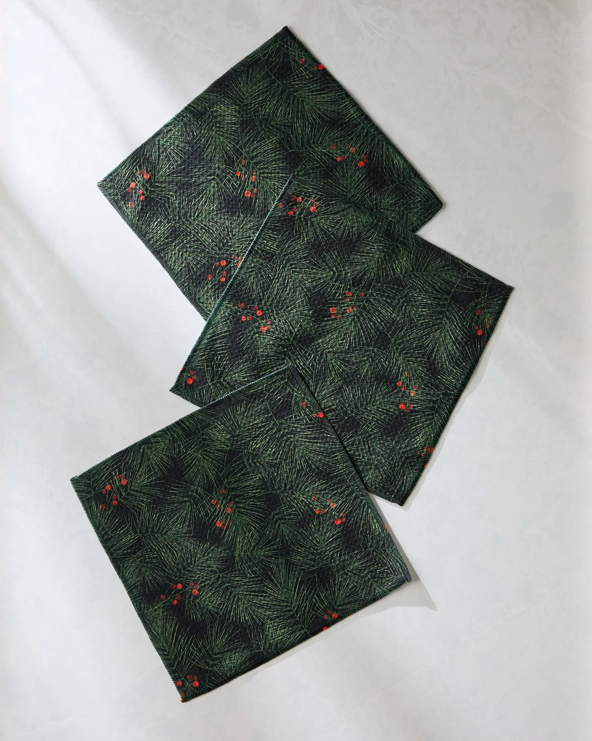 Four Evergreen Pine napkins by Geometry, featuring a green pine needle and red berry pattern, are arranged on a light surface—ideal as part of your absorbent dish towel collection for the holiday table.