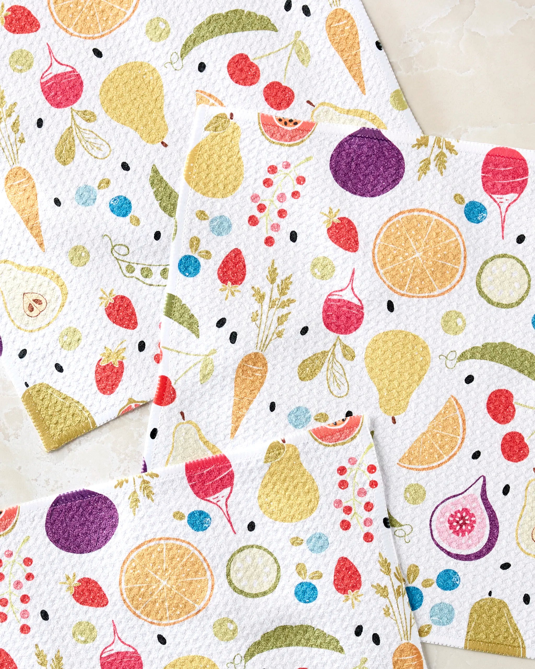 Three white dishcloths from Sara Mari's "Farmers Market" set overlap on a light surface, featuring colorful fruits and vegetables like oranges, figs, pears, peas, carrots, cherries, and black dots—made with recycled materials.