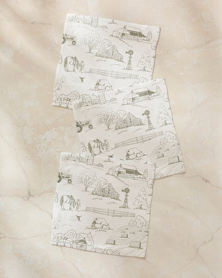 Three white "Farmyard" napkins by Hannah Langford, featuring light gray pastoral farm scenes, are overlapped on a beige marble surface—an eco-friendly dishcloth set made from recycled materials.