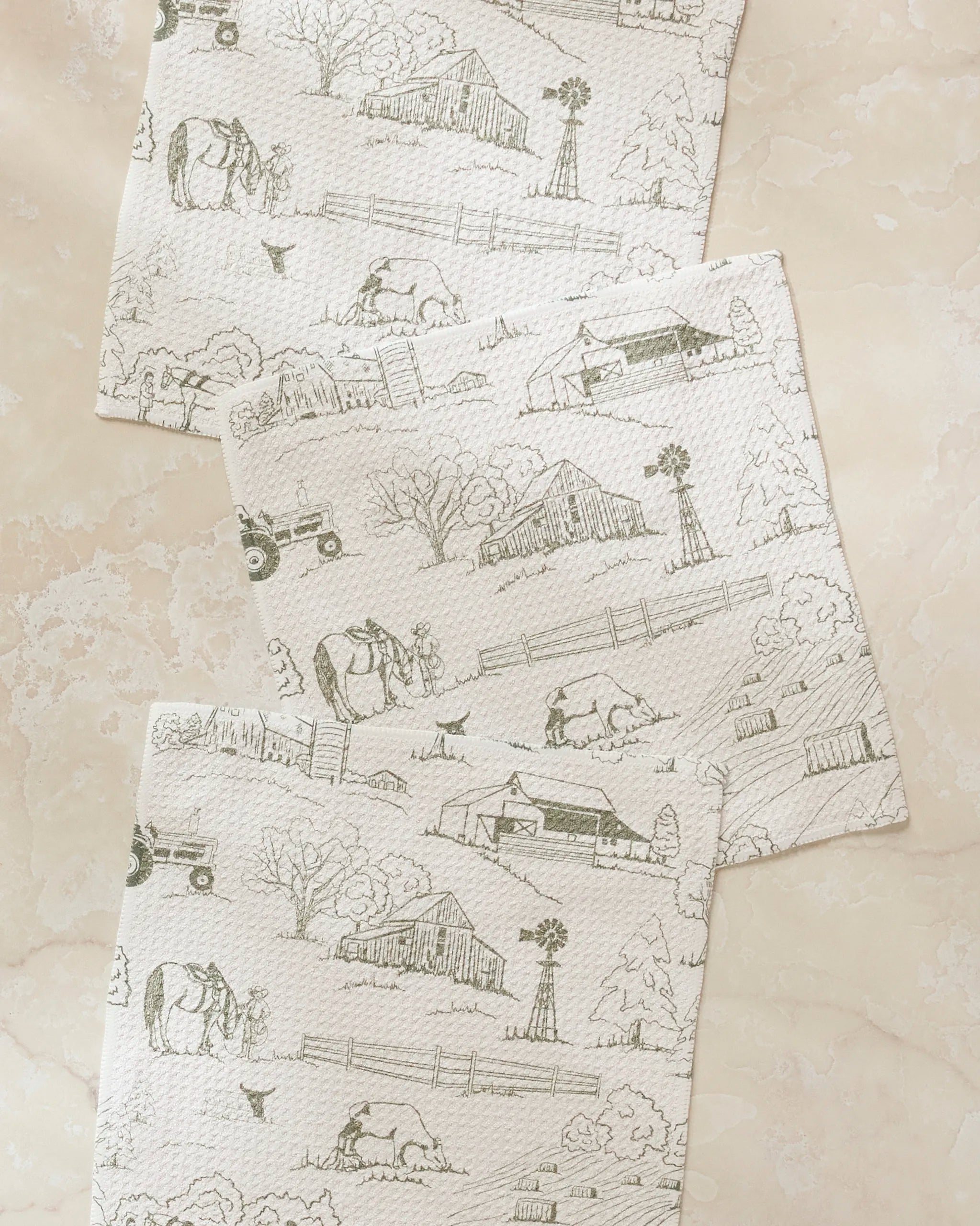 The Hannah Langford Farmyard set features three super-absorbent dishtowels with light farm scene sketches—barns, windmills, cows, and tractors—arranged diagonally on pale marble, bringing rustic charm to your kitchen routine.