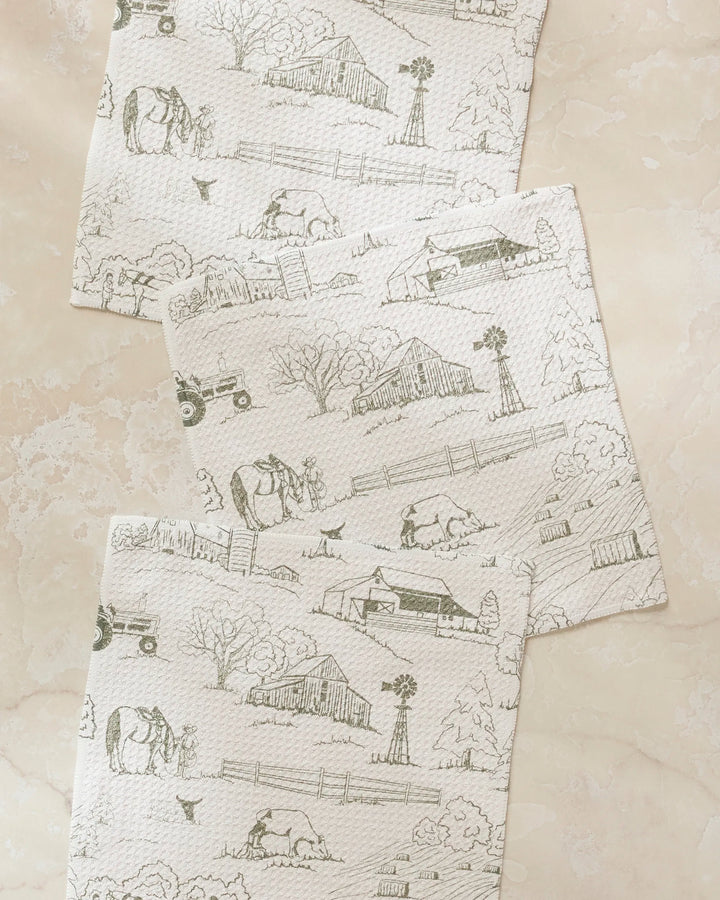 The Hannah Langford Farmyard set features three super-absorbent dishtowels with light farm scene sketches—barns, windmills, cows, and tractors—arranged diagonally on pale marble, bringing rustic charm to your kitchen routine.