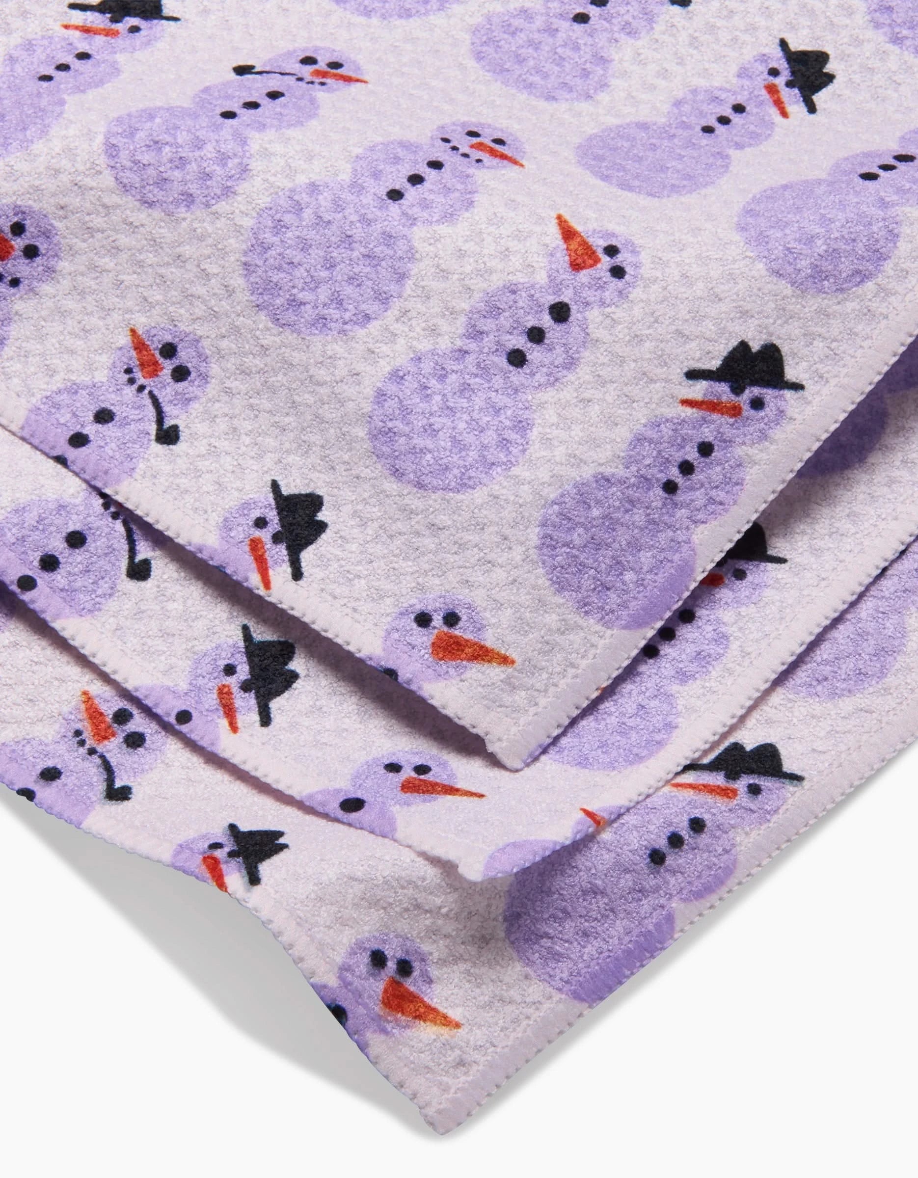 A close-up of the Amy MacCready Felicity Purple quick-drying kitchen towel, showcasing its textured fabric and repeating purple snowman pattern with orange carrot noses, black buttons, and hats on a light background.